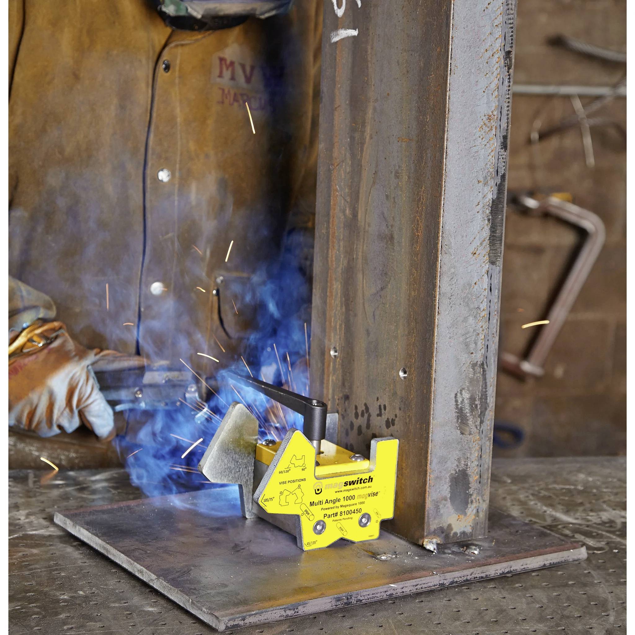 Ein Schweißer in Schutzausrüstung arbeitet an einer Metallstruktur. Eine magnetische Klemme mit der Bezeichnung 'Modell PMK-200 2000 lbs Pull 181821000' hält das Metall.