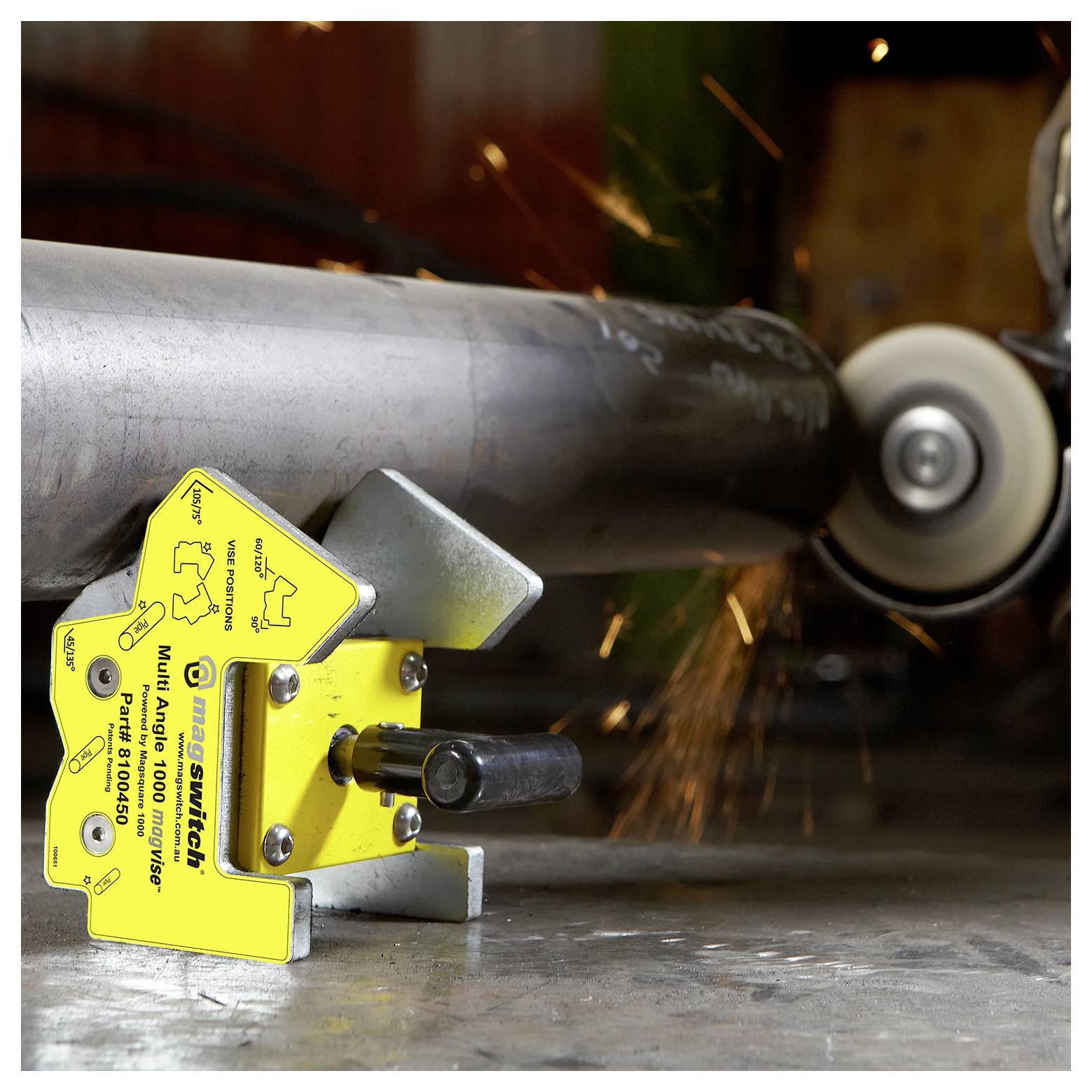 Ein Schweißmagnet hält eine Metallröhre fest, während Funken von einem Winkelschleifer aufsprühen. Der Magnet ist gelb, mit Winkeln und Sicherheitshinweisen markiert.