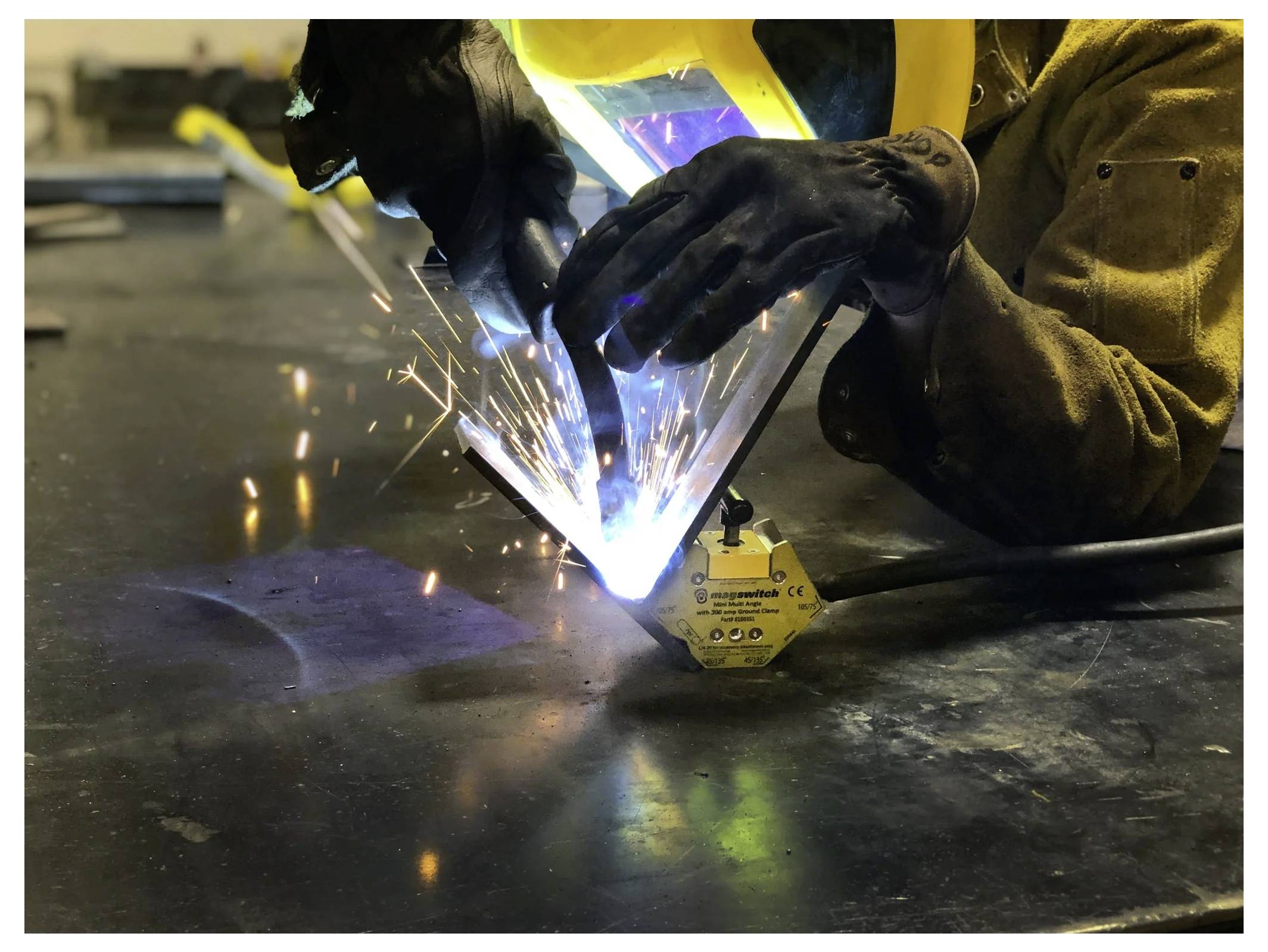 Eine Person in Schutzhandschuhen und Helm schweißt Metallteile auf einer Werkbank zusammen, wobei helle Funken die Szene beleuchten.