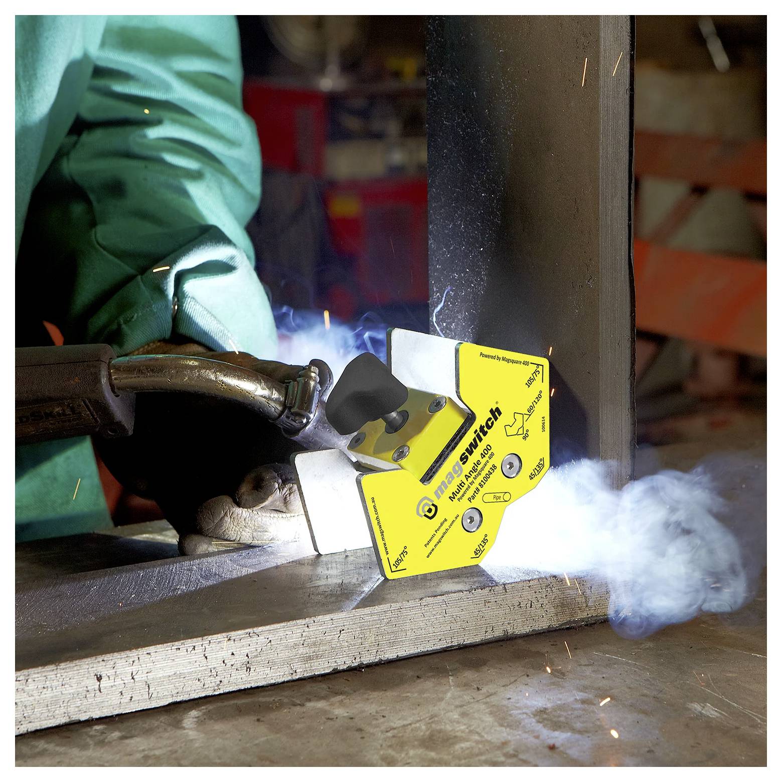 Eine Person in Schutzausrüstung schweißt Metall und verwendet einen gelben Magnethalter, um die Teile zu stützen. Rauch steigt von der Schweißtätigkeit auf.