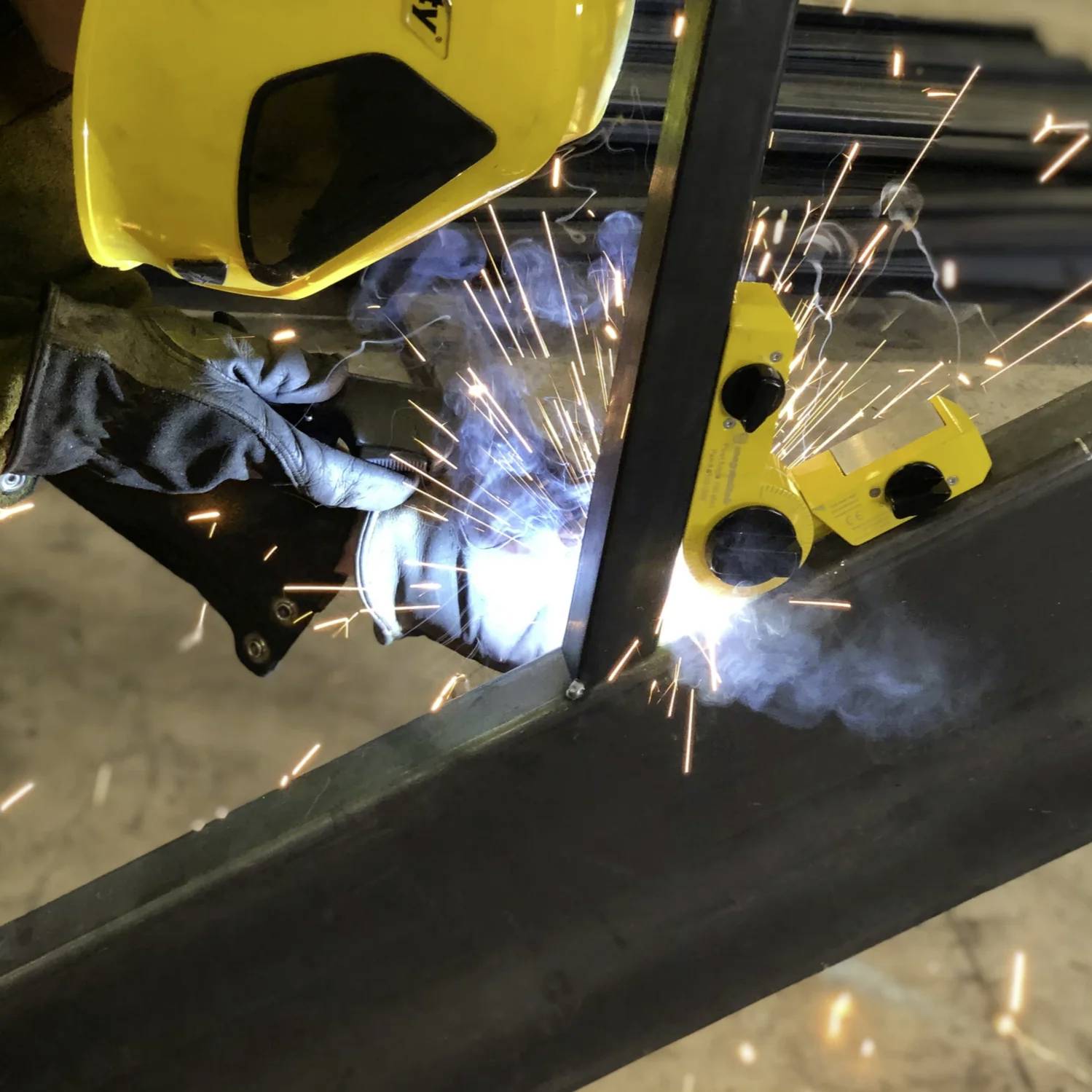 Schweißer mit gelbem Helm und Handschuhen bei der Arbeit, mit umherfliegenden Funken, der ein Schweißwerkzeug an einem Metallstück hält, in Innenräumen.