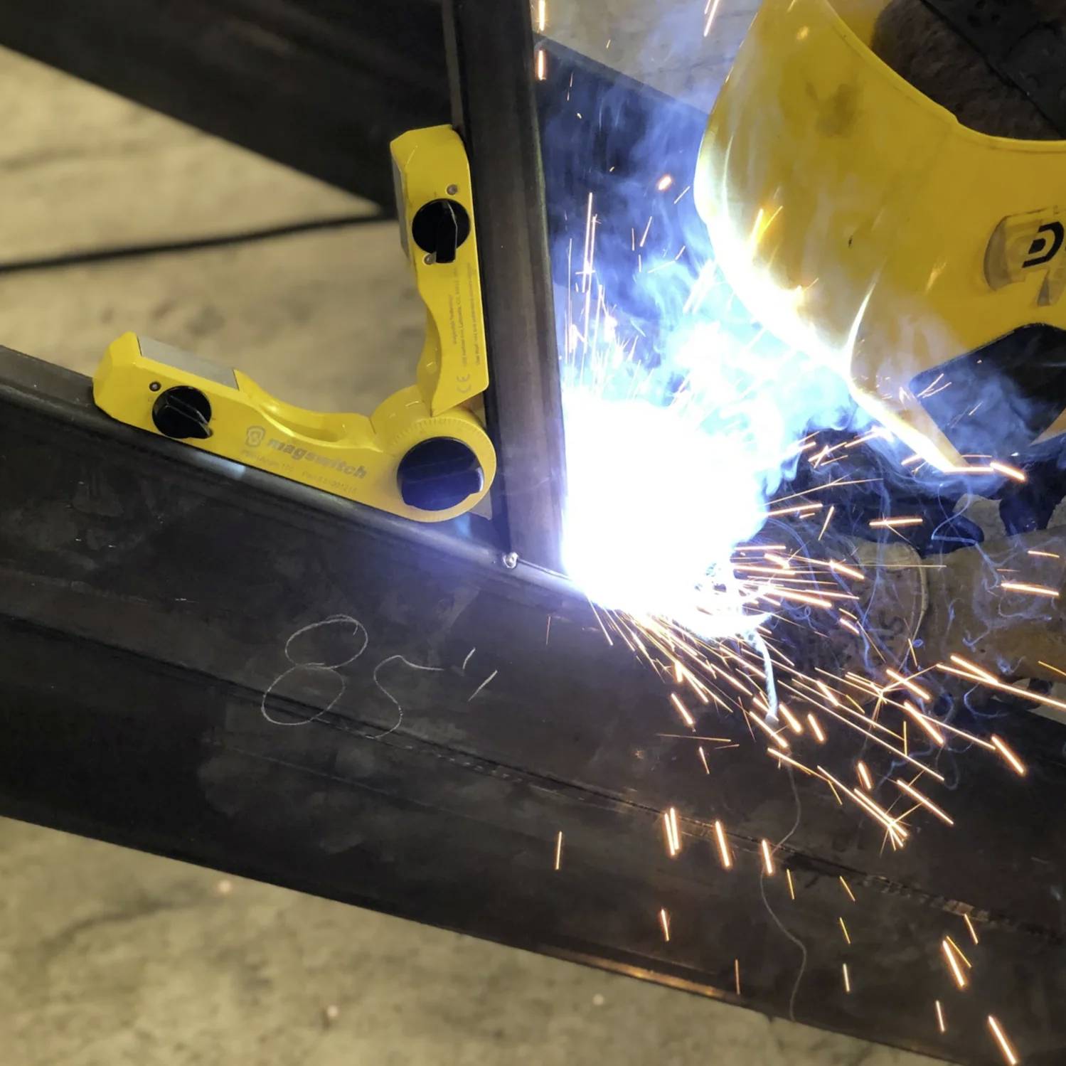 Eine Person mit einem gelben Schweißhelm arbeitet an einem Metallträger, wobei helle Schweißfunken und ein Schutzschraubstock zu sehen sind.