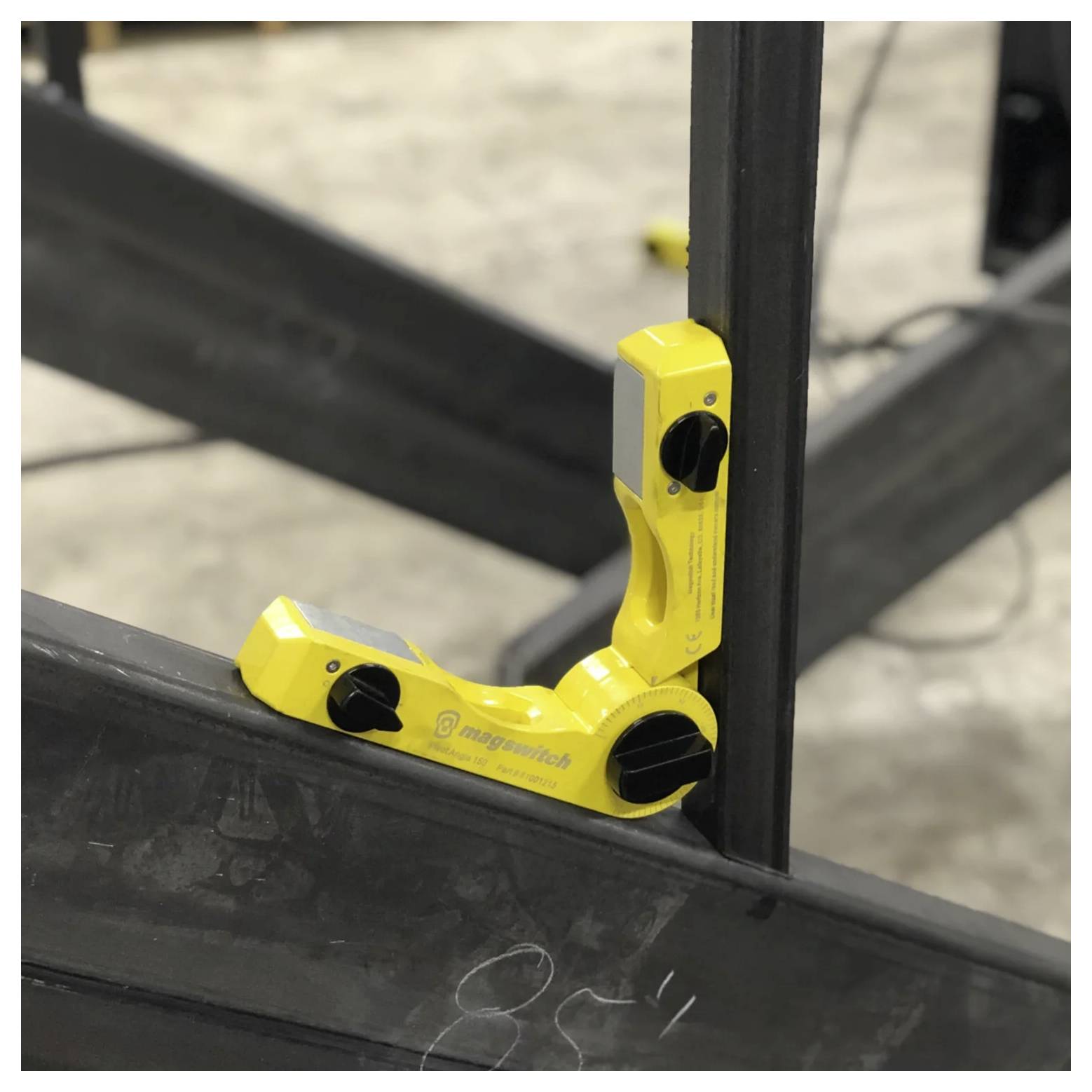 Gelbes magnetisches Werkzeug, das zwei Metallträger in einem rechten Winkel in einer Werkstatt sichert und für präzise Ausrichtung und Schweißanwendungen verwendet wird.