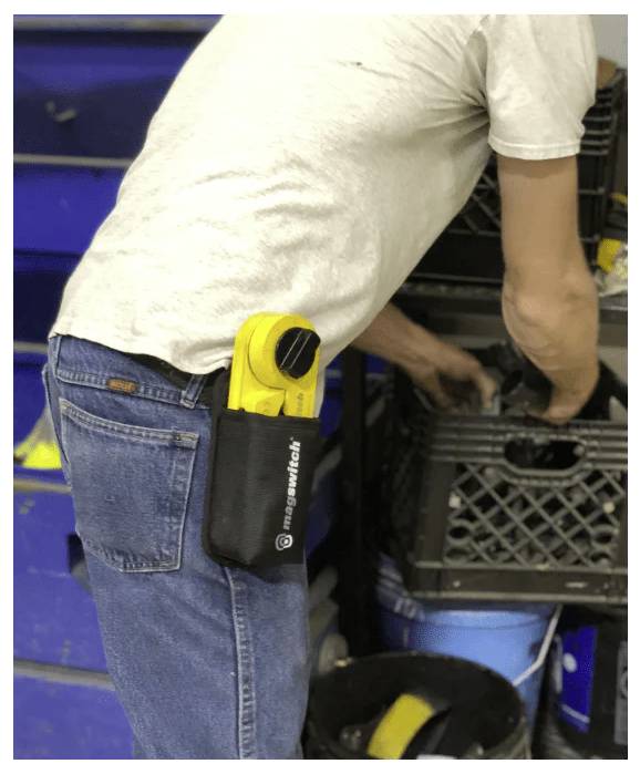 Eine Person trägt Jeans und ein weißes Hemd und sortiert Gegenstände in einer schwarzen Kiste. Ein gelbes Werkzeug ist an ihrem Gürtel befestigt.