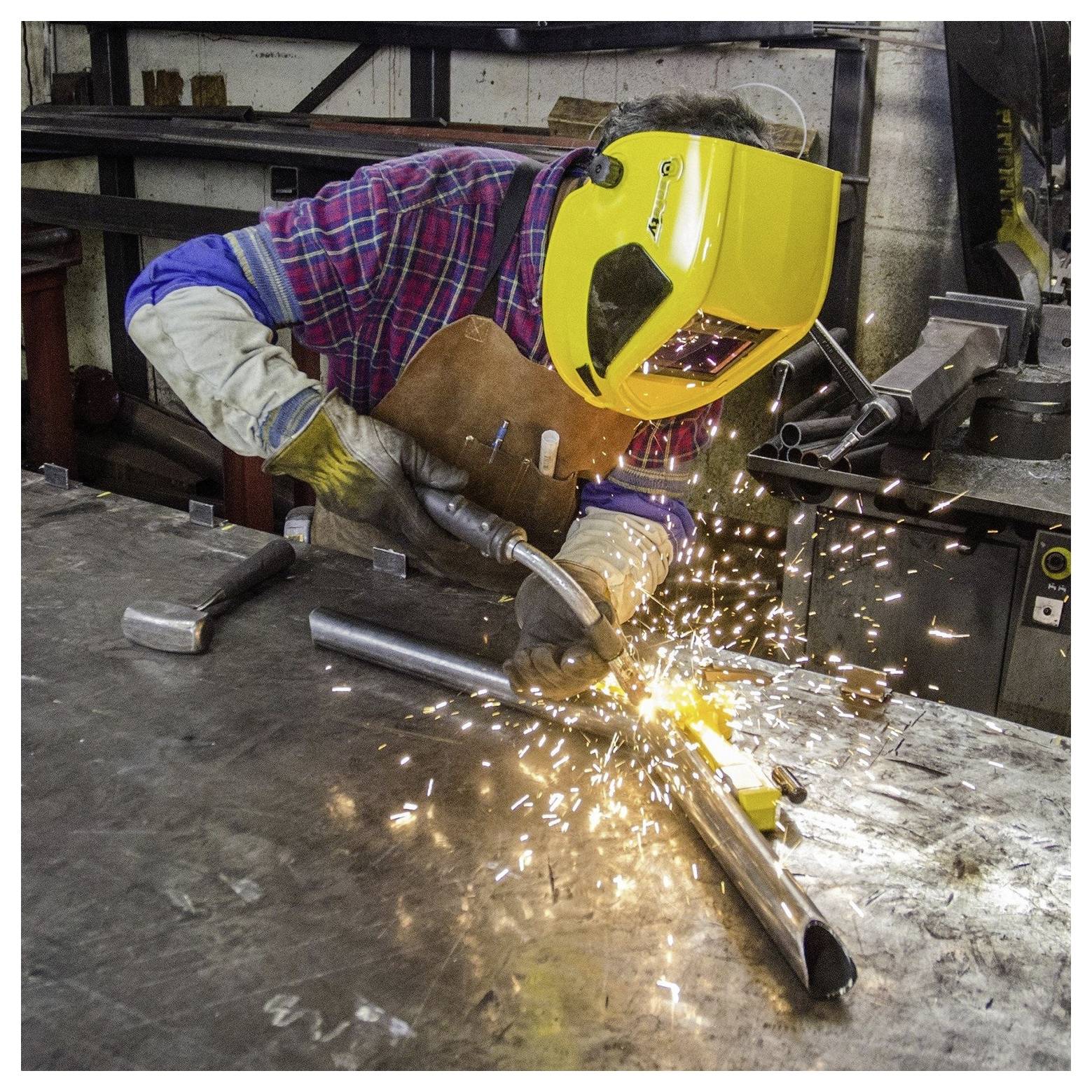 Eine Person mit Schweißmaske und Schutzausrüstung schweißt Metallteile auf einer Werkbank und erzeugt dabei helle Funken in einer industriellen Umgebung.