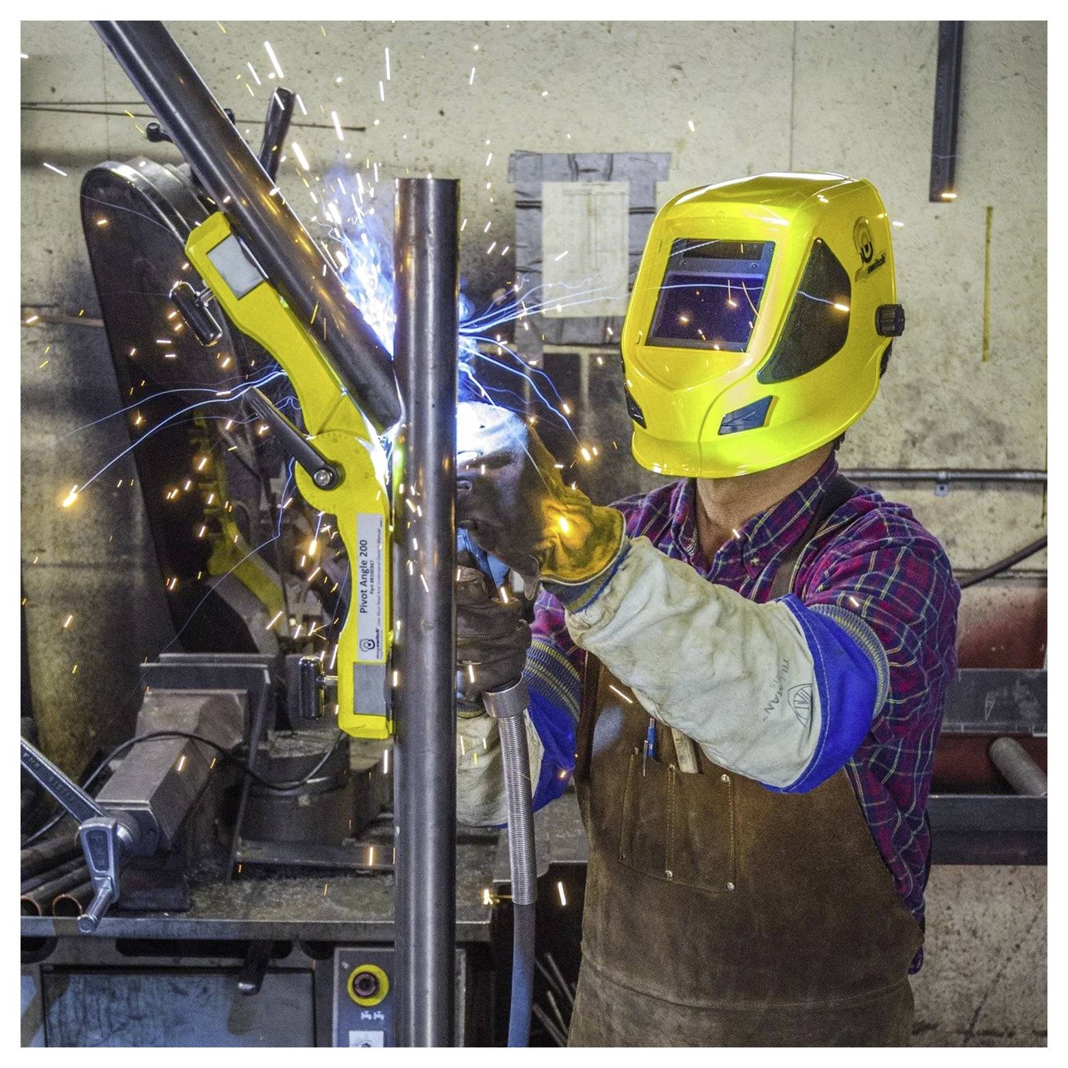 Eine Person mit gelbem Schweißhelm und Schutzausrüstung schweißt ein Metallrohr, wobei Funken in einer Werkstattumgebung sichtbar sind.
