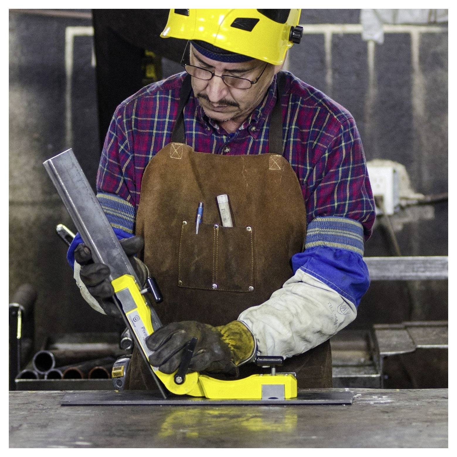 Eine Person in Sicherheitsausrüstung, einschließlich eines gelben Helms und einer braunen Schürze, verwendet ein Messwerkzeug, um die Ausrichtung an einem Metallstück in einer Werkstatt zu überprüfen.