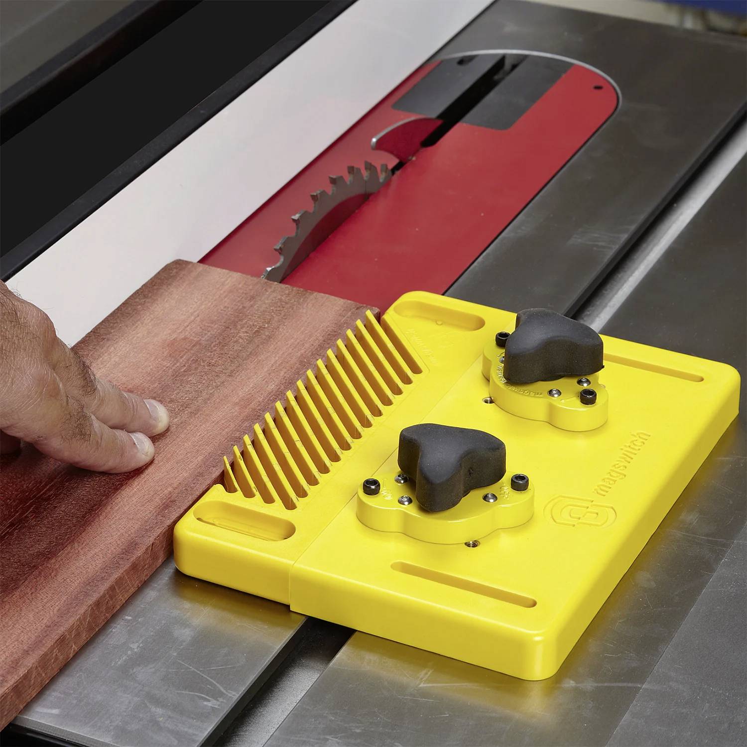 Eine Hand führt ein Holzbrett durch eine Kreissäge und verwendet dabei ein gelbes Sicherheitswerkzeug, um die Finger vom Sägeblatt fernzuhalten.