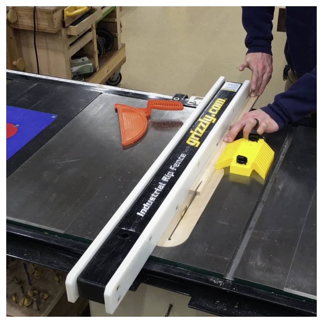 Eine Person führt ein Stück Holz durch eine Kreissäge mit einer mit der Marke 'grizzly.com' versehenen Führungsschiene und verwendet dabei einen gelben Sicherheits-Schiebeholz.