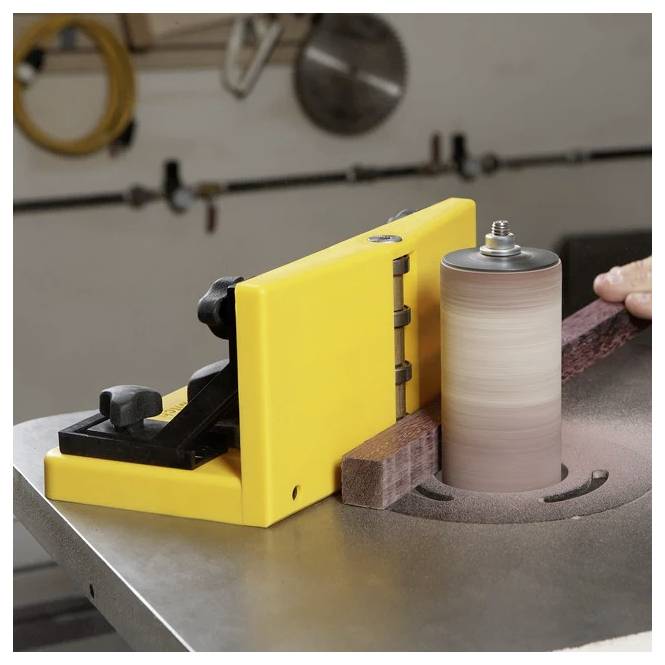 Eine Spindelschleifmaschine auf einer Werkbank mit einer gelben Sicherheitsabdeckung. Ein Holzbrett wird geschliffen, wobei sich feine Sägespäne um die Basis herum bilden.