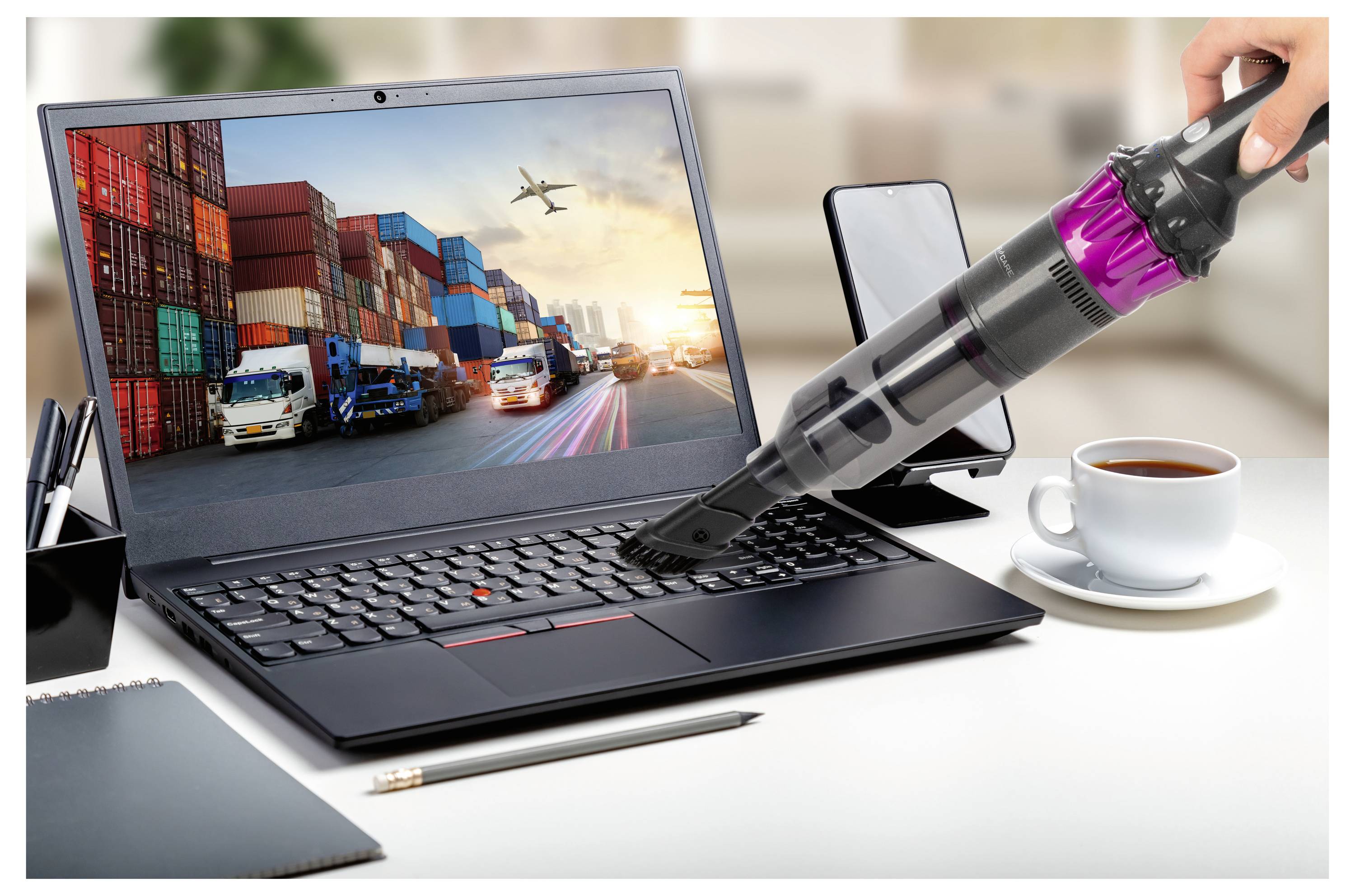 Ein Laptop zeigt eine Frachtversandansicht, während ein Mini-Staubsauger seine Tastatur reinigt. In der Nähe befinden sich ein Smartphone, ein Stift, ein Notizblock und eine Kaffeetasse.