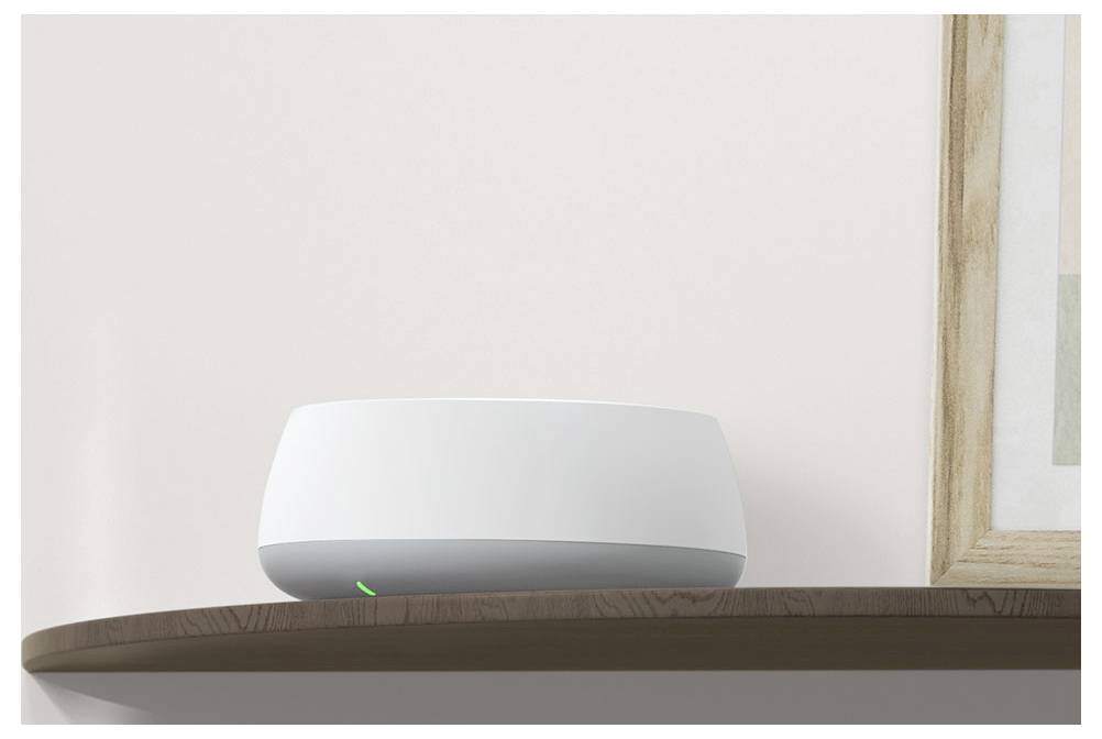 Ein moderner weißer Router mit einem leuchtenden grünen Licht steht auf einem braunen Regal vor einer schlichten Wand, in der Nähe eines Bildes mit einem Holzrahmen.