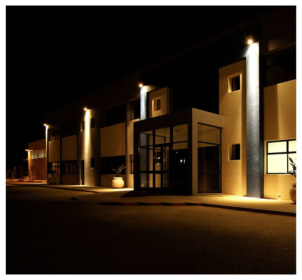 Ein modernes Gebäude bei Nacht mit beleuchtetem Eingang und Außenwandleuchten, die warmes Licht auf die Fassade werfen.