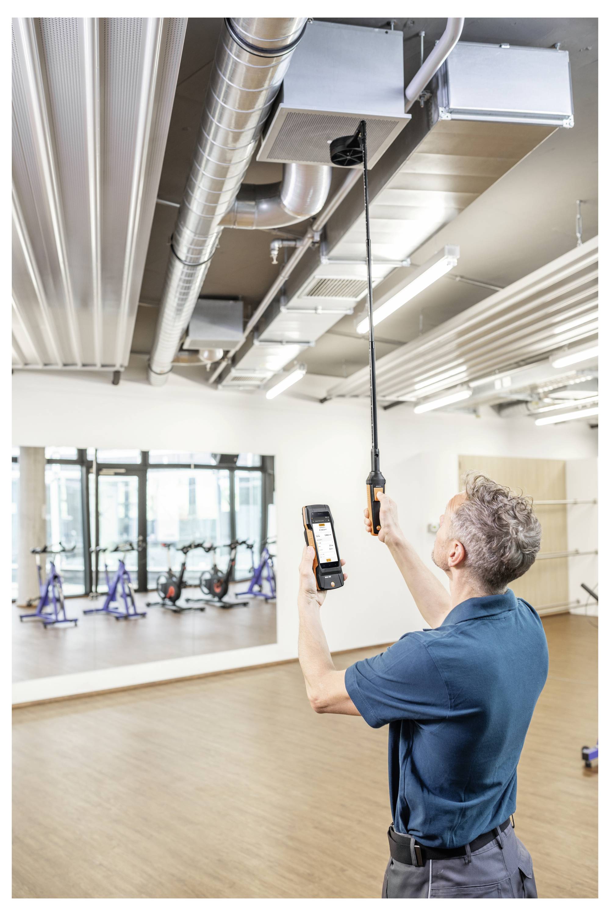 Eine Person in einem blauen Hemd inspiziert Lüftungskanäle in einem Fitnessstudio und verwendet dabei ein Handgerät und ein Smartphone zur Messung.