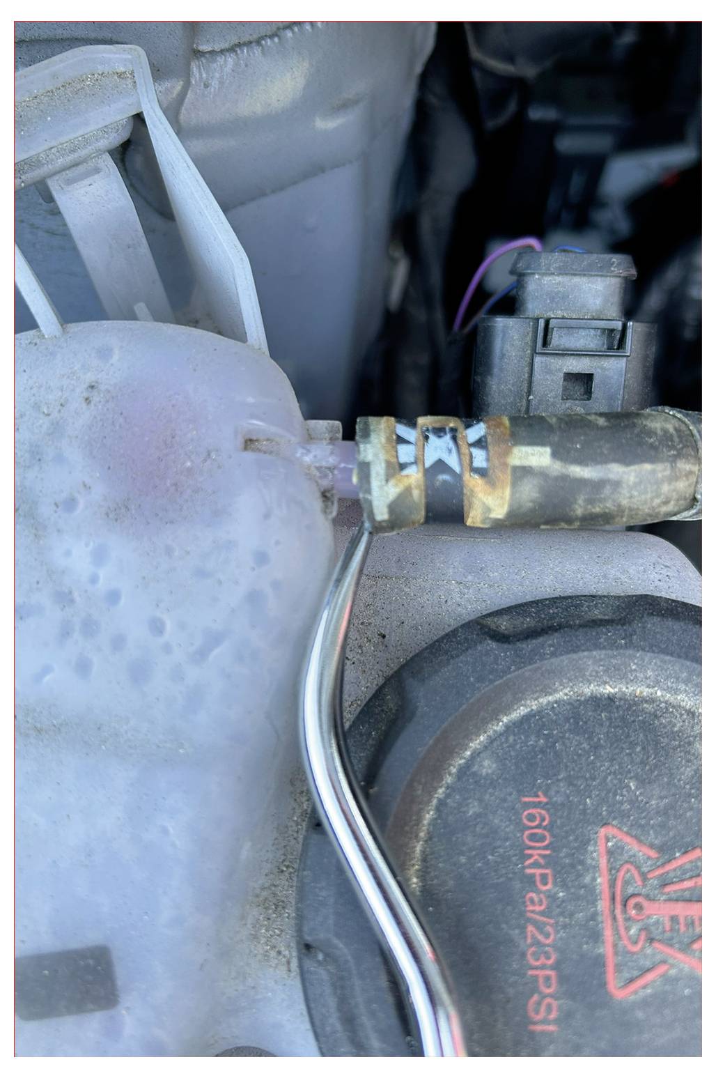 Eine Nahaufnahme einer Motorkomponente, die eine rostige Metallklemme an einem Schlauch zeigt, der mit einem weißen Plastikbehälter verbunden ist. Ein Deckel ist mit '160KPa/23PSI' beschriftet.