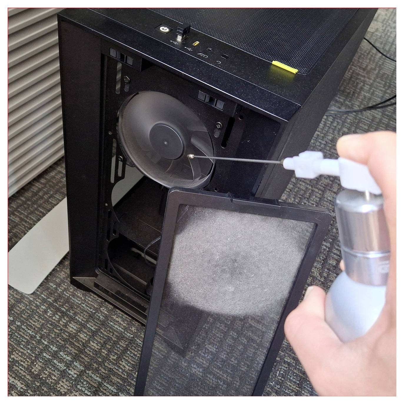 Eine Person verwendet Druckluft, um einen Computerlüfter innerhalb eines Gehäuses zu reinigen, wobei der Staubfilter entfernt und das Gehäuse auf dem Boden steht.