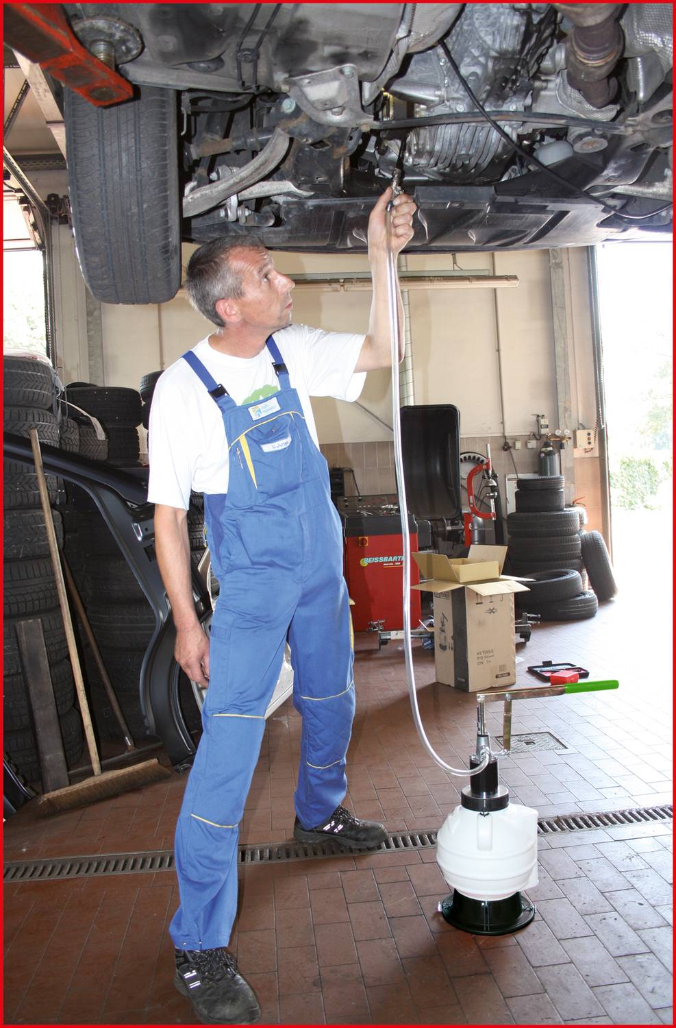 Ein Mechaniker in blauen Overalls wechselt das Öl eines angehobenen Fahrzeugs mit Hilfe eines Schlauchs und einer Pumpe in einer Autowerkstatt, umgeben von Werkzeugen und Reifen.