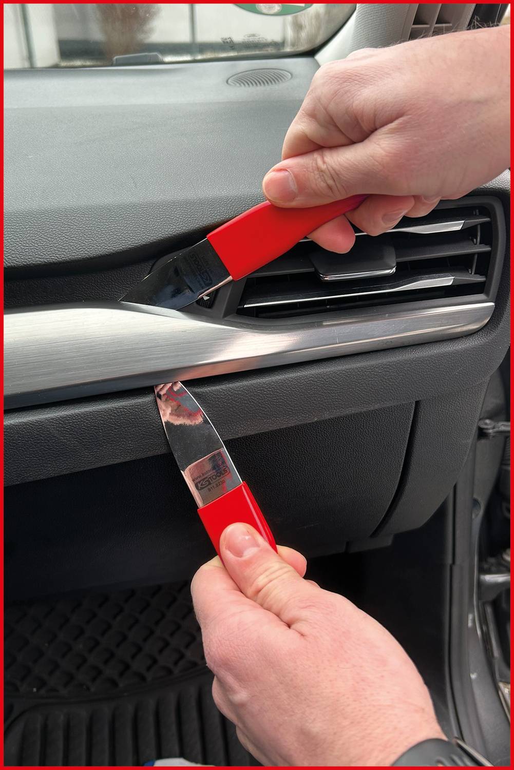 Eine Person verwendet zwei rote Kunststoffwerkzeuge, um eine Innenverkleidung im Fahrzeug in der Nähe der Lüftungsöffnung zu entfernen und zeigt dabei einen DIY-Reparaturprozess.