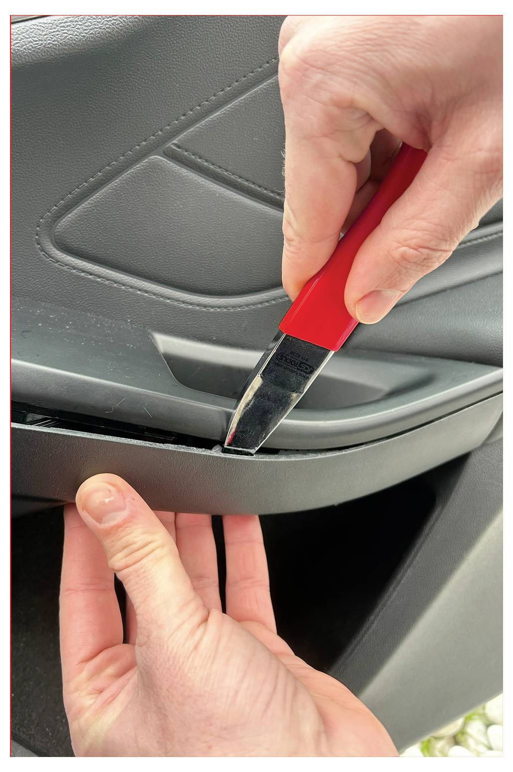 Zwei Hände benutzen ein rotgriffiges Werkzeug, um vorsichtig das Innenverkleidungspanel einer Autotür zu öffnen und dabei eine Reparatur- oder Wartungsaufgabe durchzuführen.