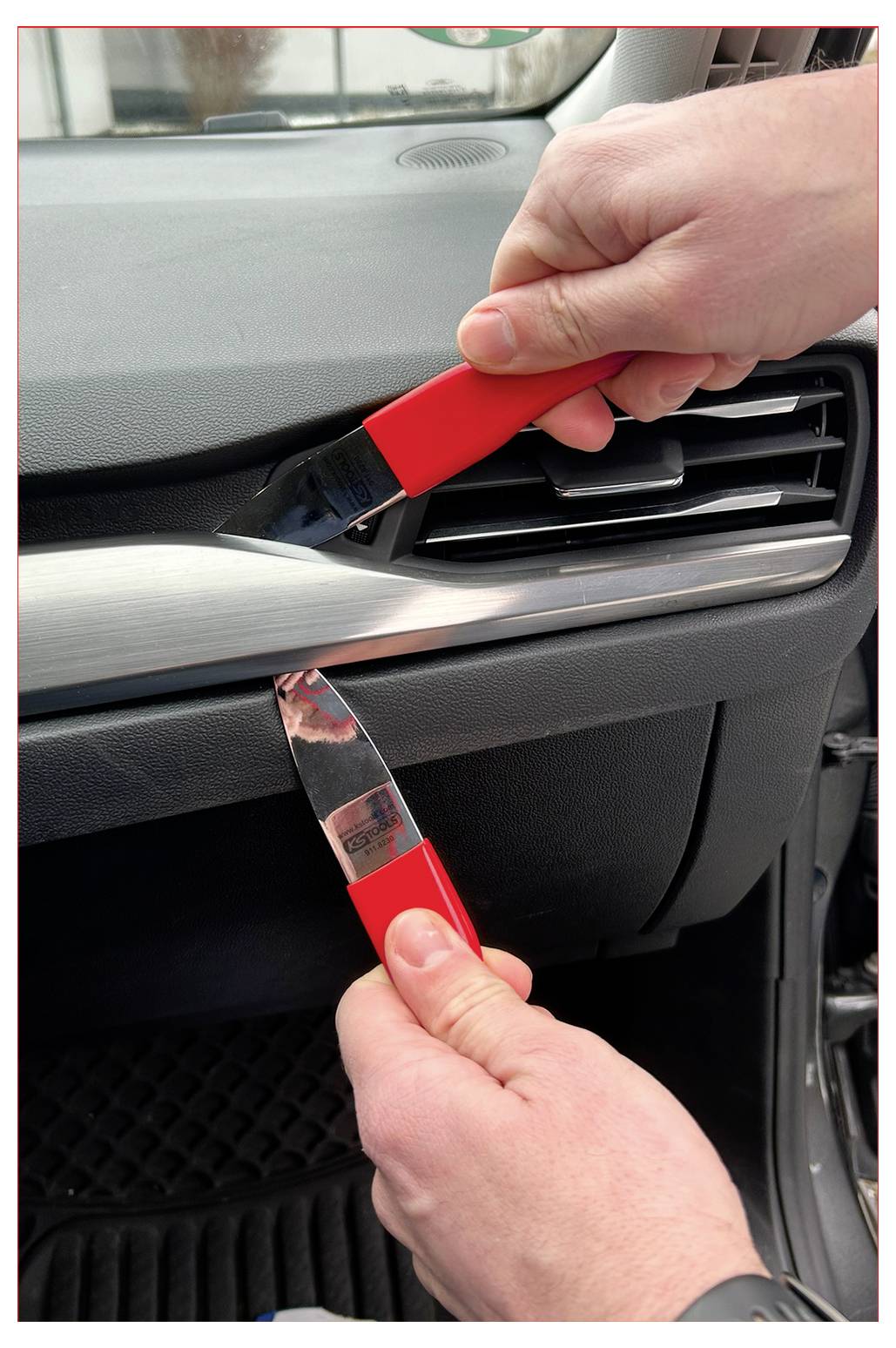 Zwei Hände, die rote Plastikwerkzeuge verwenden, um eine Innenverkleidung eines Autos zu entfernen und dabei eine Lüftungsöffnung freizulegen.