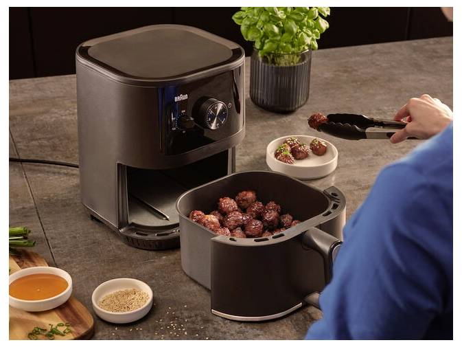 Eine Person benutzt eine Zange, um Fleischbällchen in eine Schüssel neben einem Heißluftfritteuse auf einer Küchenarbeitsplatte zu legen, wobei Gewürze und Sauce in der Nähe sind.