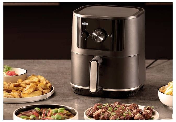 Eine Küchenarbeitsplatte mit einem schwarzen Heißluftfritteusen, umgeben von Tellern mit Pommes, Hühnerflügeln und Gemüse, die eine Mahlzeitenzubereitungsszene andeuten.