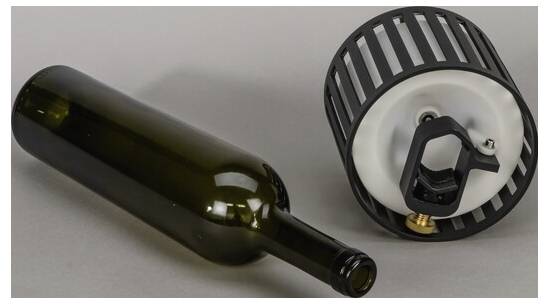 Eine Weinflasche liegt neben einem modernen, zylindrischen Objekt in Schwarz und Weiß, möglicherweise ein Haushaltsgerät oder Geräteteils, auf einer grauen Oberfläche.