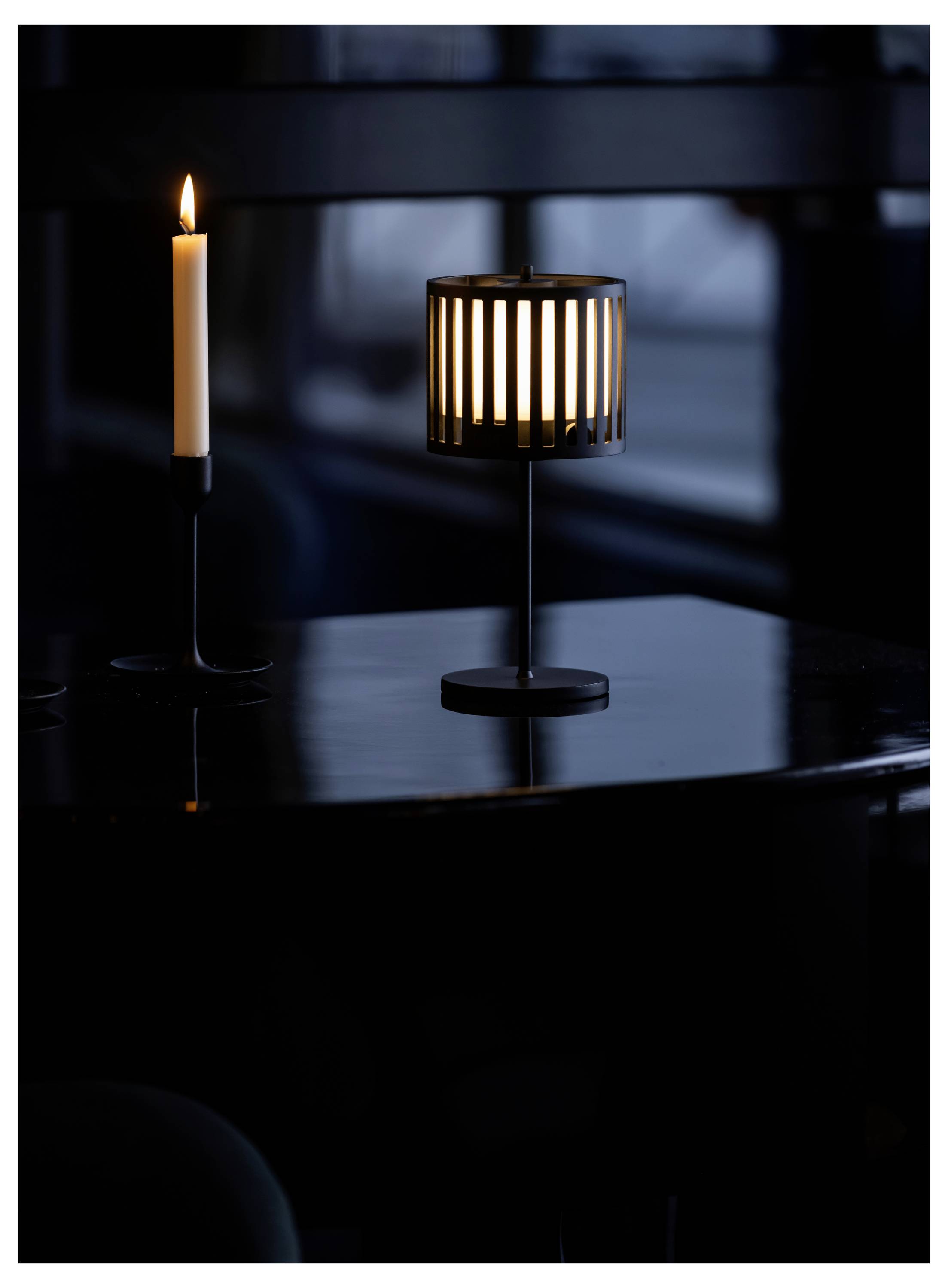 Eine brennende Kerze und eine Lampe mit vertikal gestreiftem Lampenschirm auf einem dunklen Tisch, die sanftes Licht spenden, vor einem dunkel beleuchteten Hintergrund.