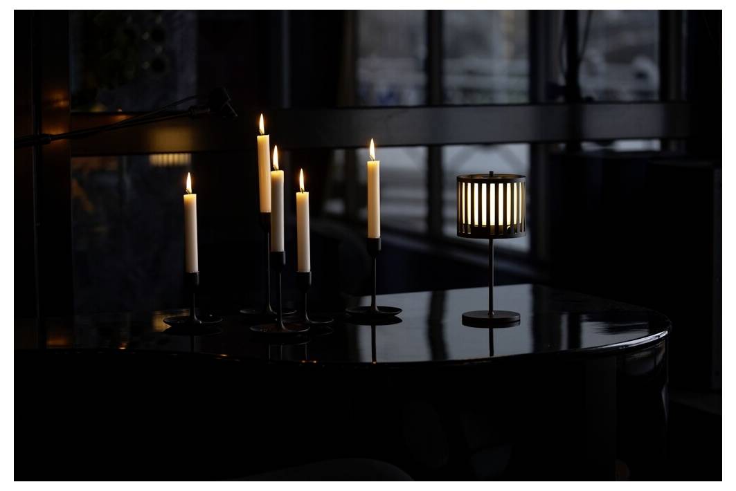 Kerzen auf einem dunklen Klavier angezündet, die weiches Licht reflektieren; eine moderne Lampe sorgt für gedämpftes Ambiente, aufgestellt in einem dämmrigen Raum mit großen Fenstern.