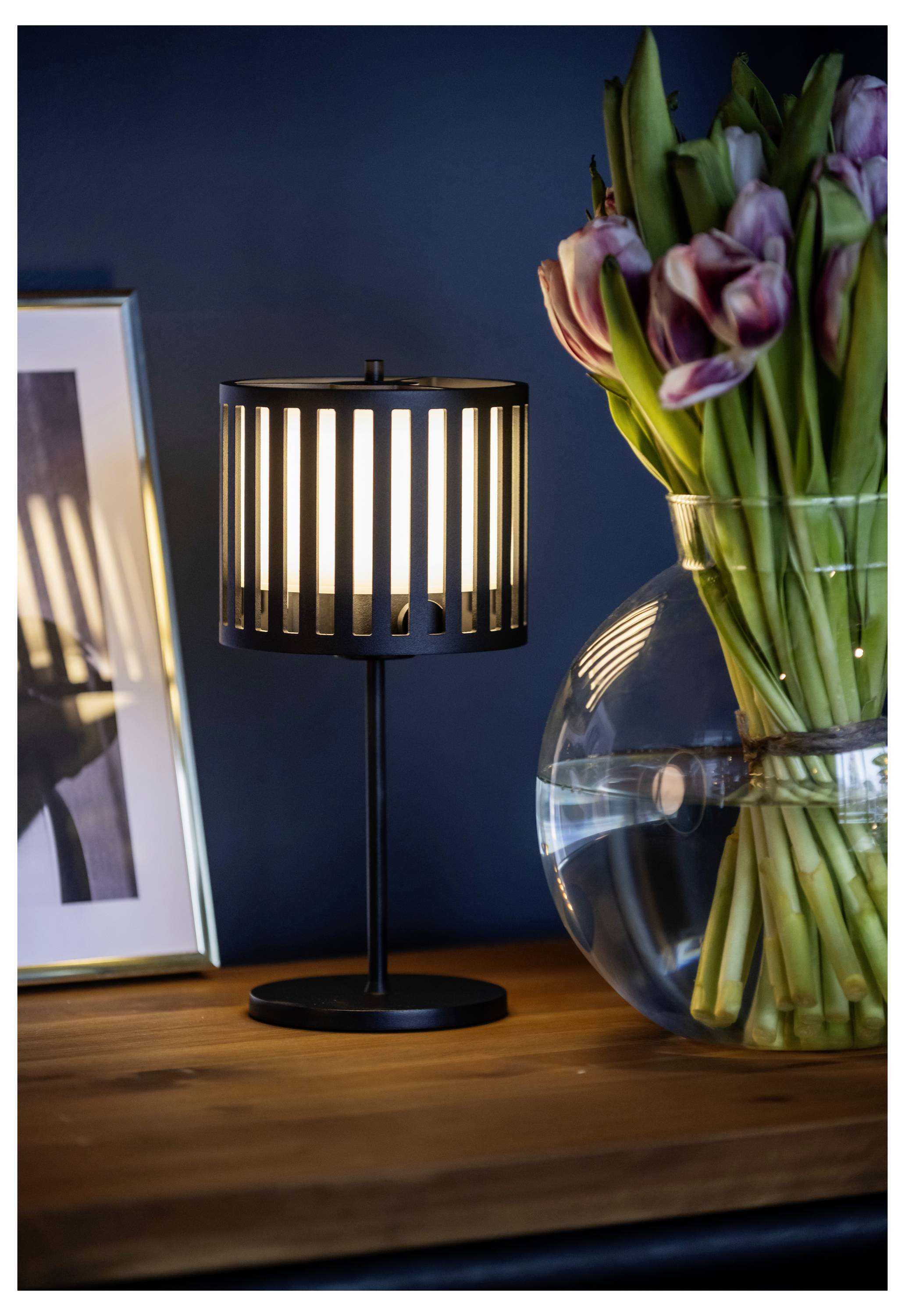 Eine warm beleuchtete schwarze Tischlampe und eine Vase mit rosa Tulpen stehen auf einem Holztisch, neben einem gerahmten Foto, vor einer dunkelblauer Wand.