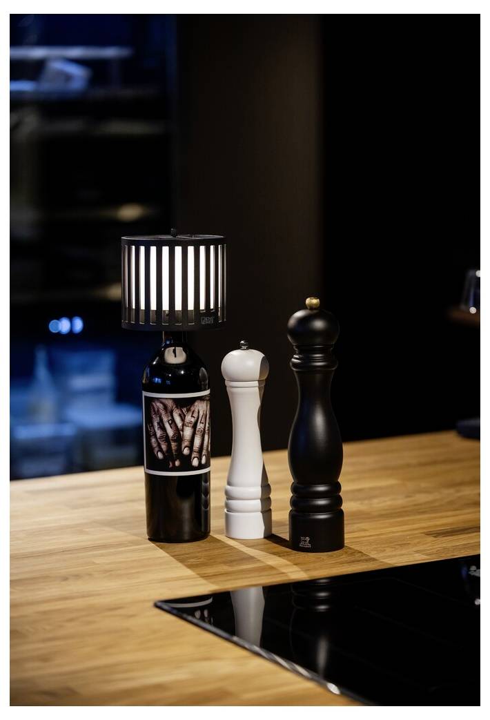 Eine Flasche mit einer kleinen Lampe daran steht auf einer Holztheke, neben weißen und schwarzen Pfeffermühlen. Der Hintergrund ist sanft beleuchtet.