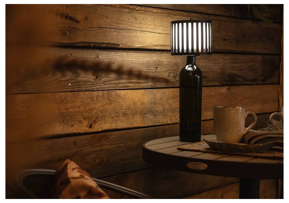 Ein gemütlicher, dämmriger Holzraum mit einem Tisch, auf dem eine Tasse, Untertasse und eine Lampe stehen, die aus einer Flasche gefertigt ist und gestreifte Schatten an die Wand wirft.