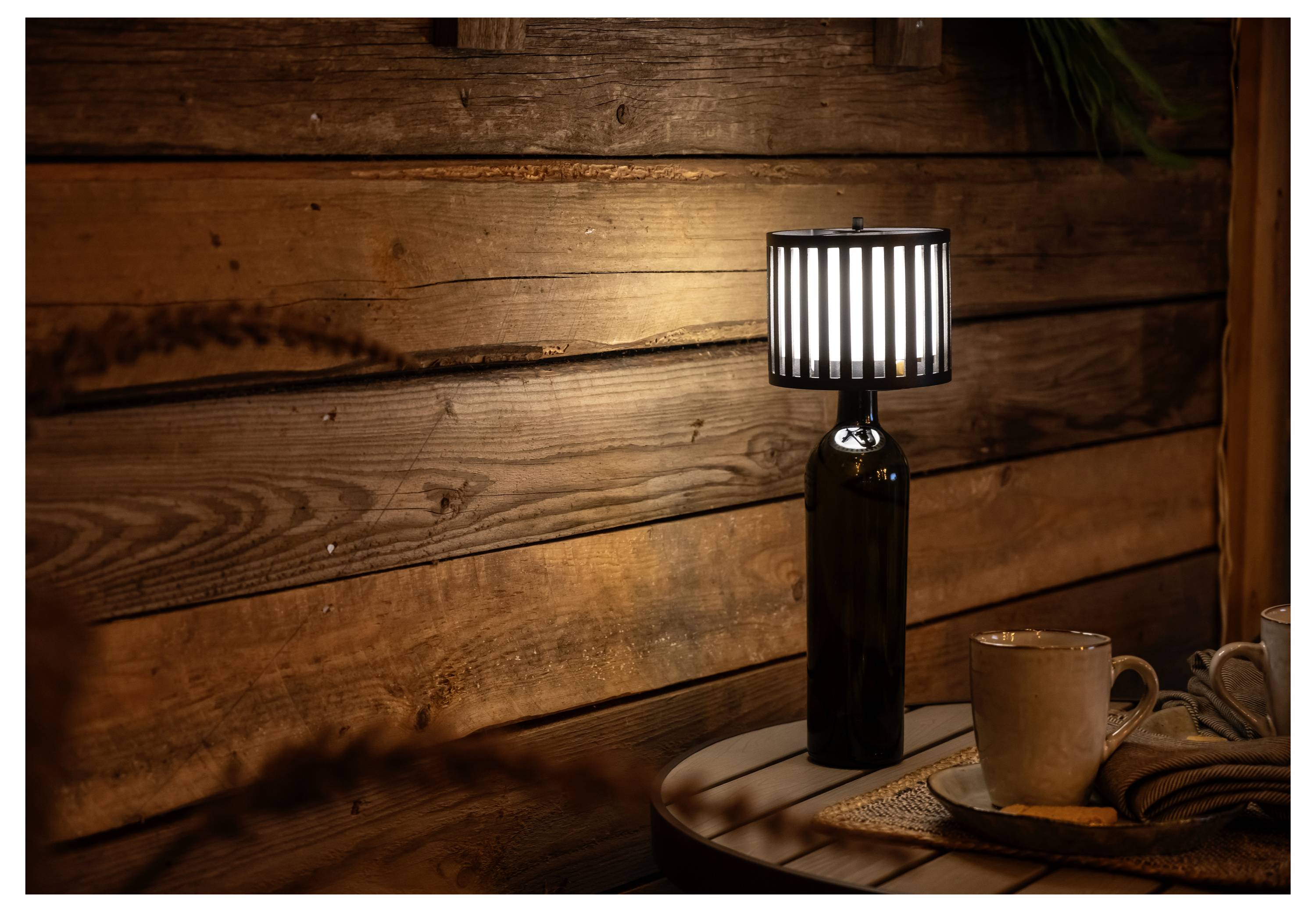 Ein gemütlicher Holzraum zeigt eine einzigartige Lampe, die aus einer Glasflasche mit einem gestreiften Lampenschirm auf einem runden Tisch besteht, neben Kaffeetassen und Weizen.