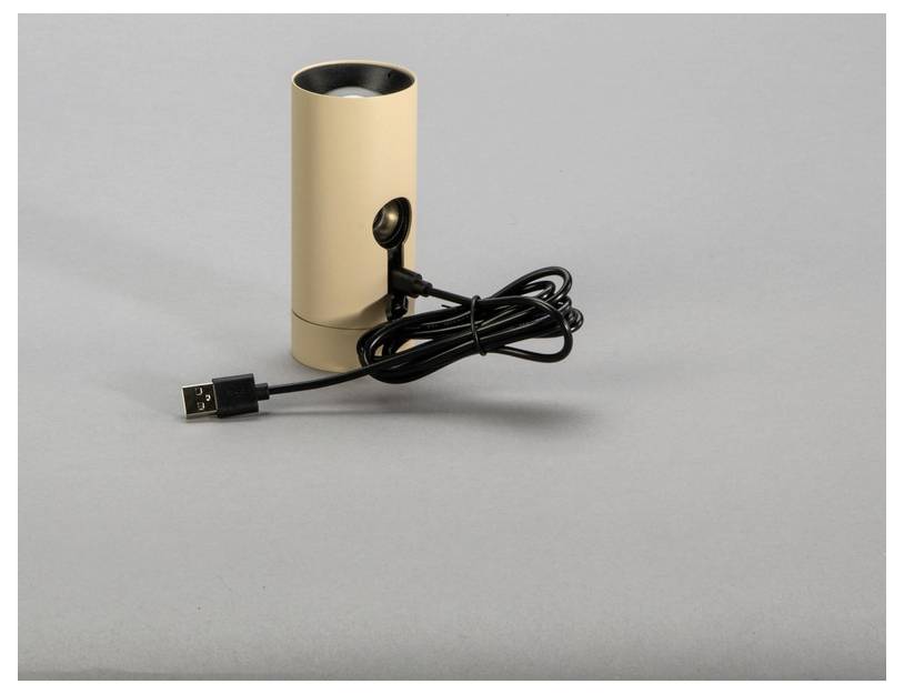 Ein beiger zylindrischer Lautsprecher mit einem USB-Kabel liegt auf einer grauen Oberfläche.