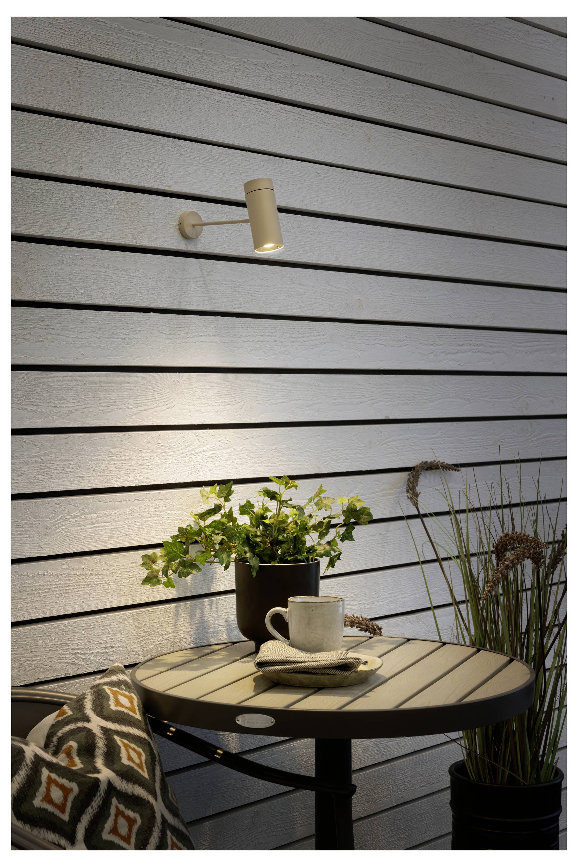 Kleine Terrasseneinrichtung mit einem runden Tisch, auf dem eine Tasse und Untertasse stehen. Eine Pflanze und Gräser daneben, beleuchtet von einer wandmontieren Leuchte.