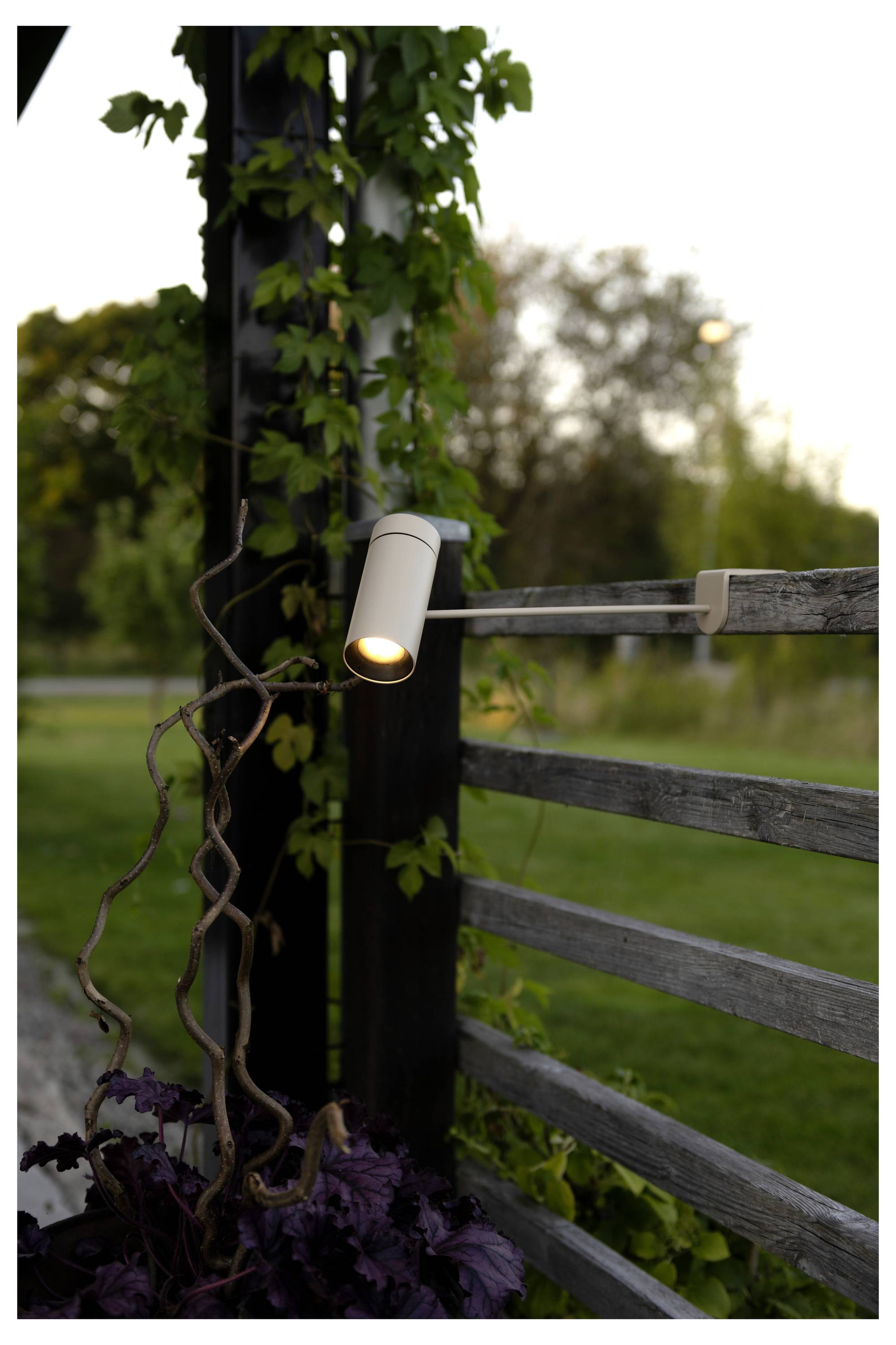 Eine kleine Außenleuchte beleuchtet einen Holzzaun und kletternde Pflanzen in der Abenddämmerung und schafft eine friedvolle Gartenatmosphäre.