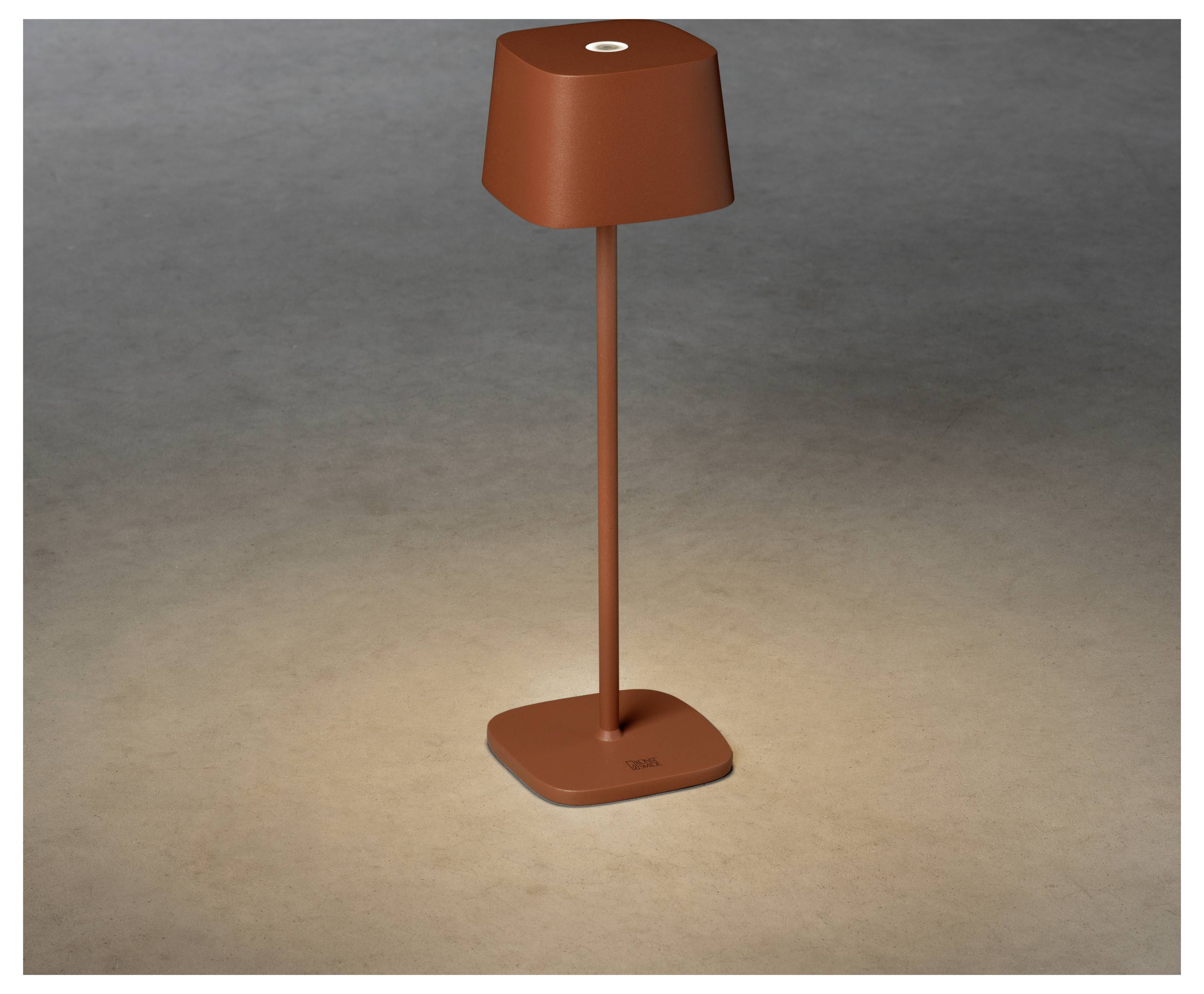 Eine elegante, moderne Tischlampe mit quadratischer Basis in einer rot-braunen Farbe, die die Oberfläche darunter in einem dezenten, warmen Licht beleuchtet.