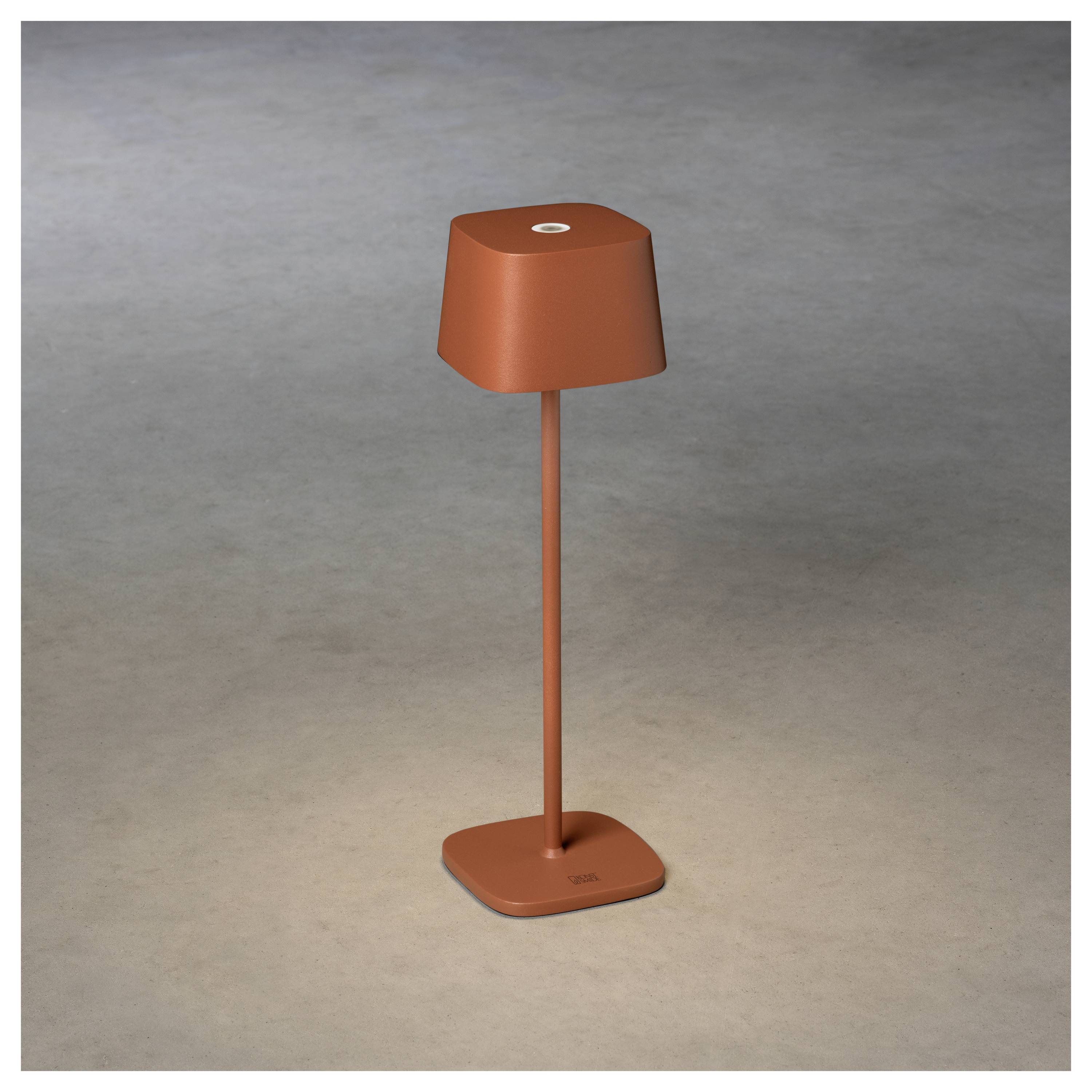 Eine minimalistische rote Tischlampe mit quadratischem Fuß und Lampenschirm, die einen weichen Schein auf neutralem Hintergrund verbreitet.