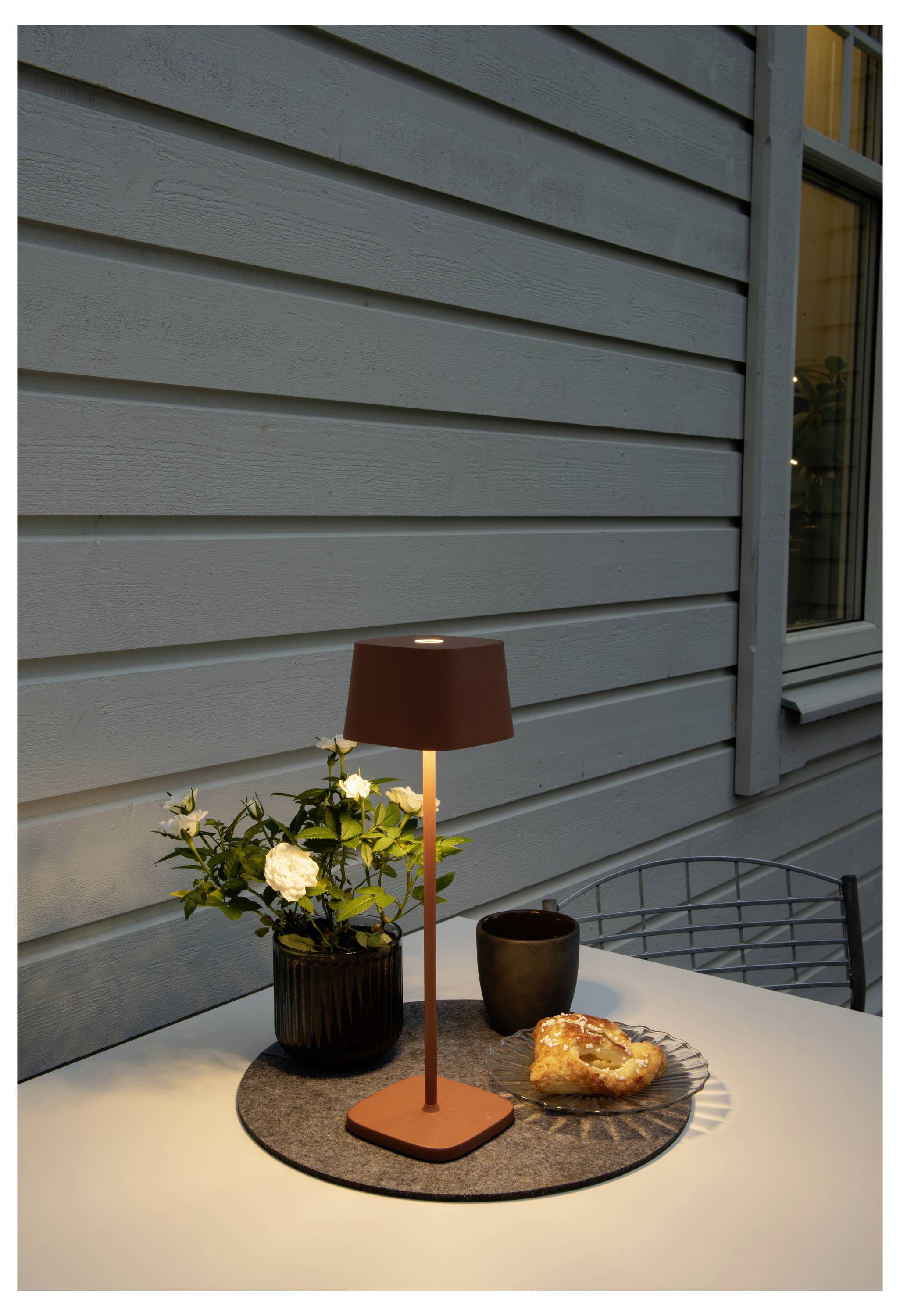 Ein runder Tisch mit einer kleinen Lampe, einer Topfpflanze, einer Tasse Kaffee und einem Gebäckstück ist im Freien vor einer Holzwand platziert.