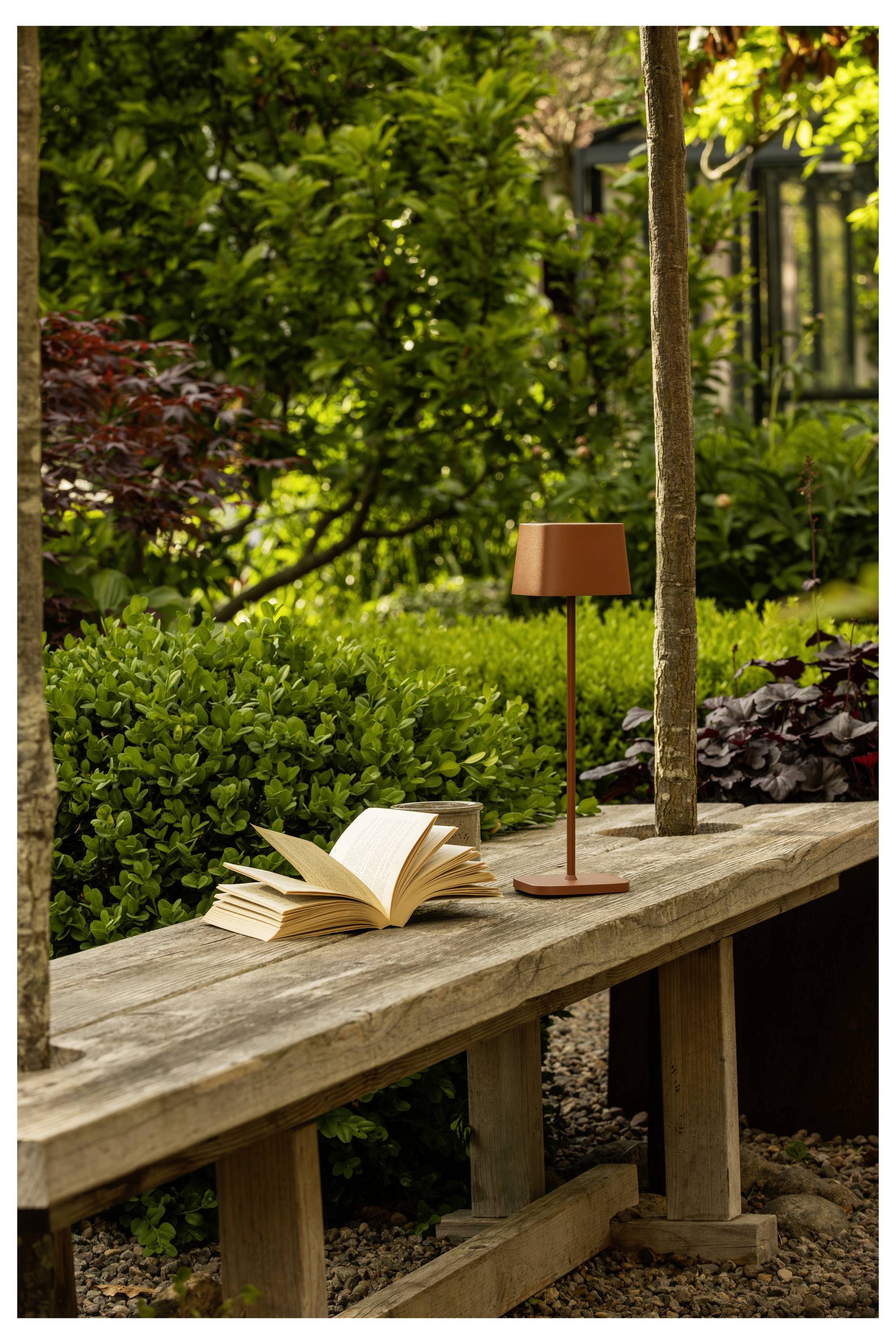 Eine Holzbank in einem Garten mit einem aufgeschlagenen Buch und einer kleinen Lampe darauf, umgeben von üppigem grünem Laub und weichem natürlichem Licht.