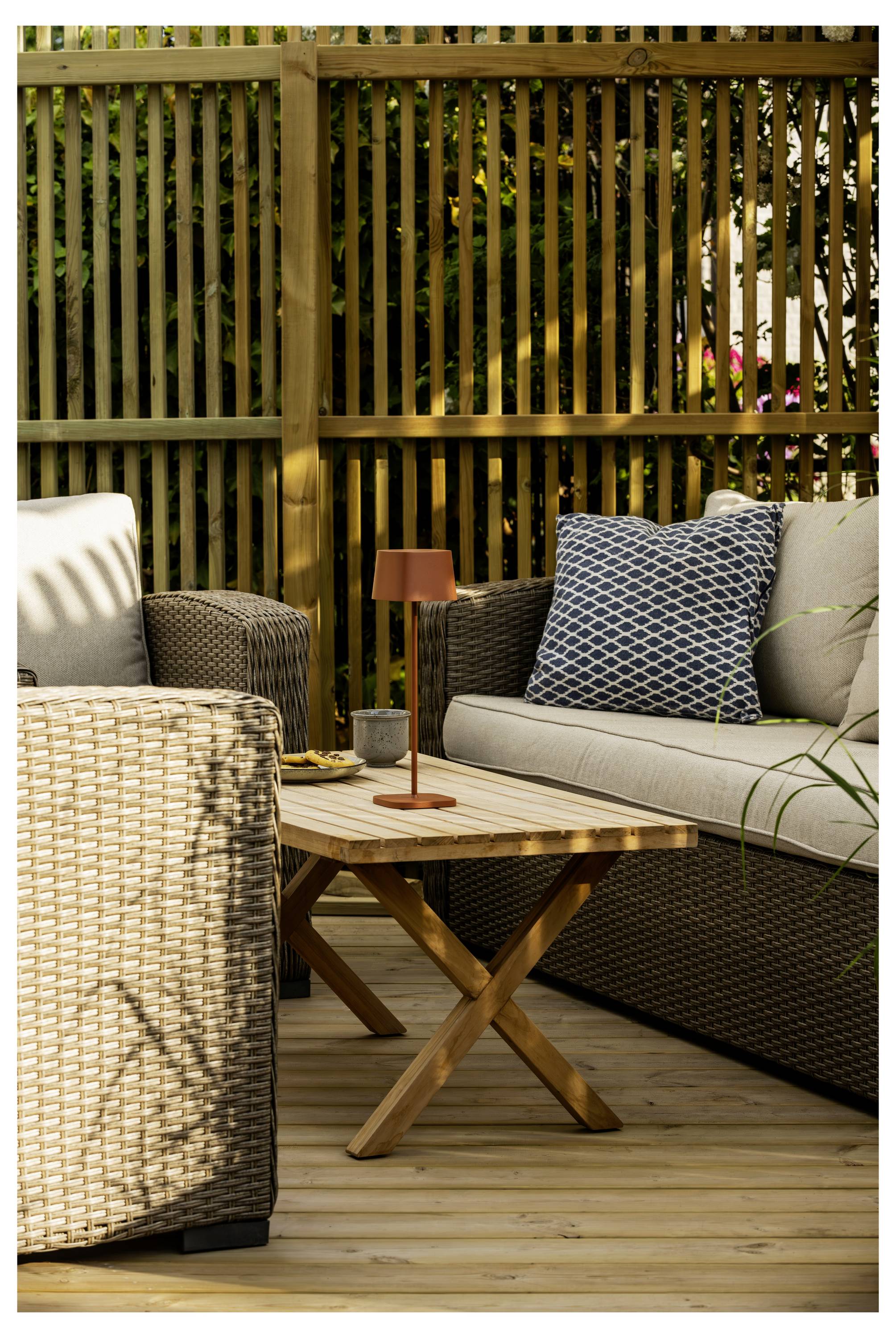 Außenbereich mit Sitzgelegenheiten, bestehend aus Rattanmöbeln, einem Holztisch und einem gemusterten Kissen in einem eingezäunten Garten.