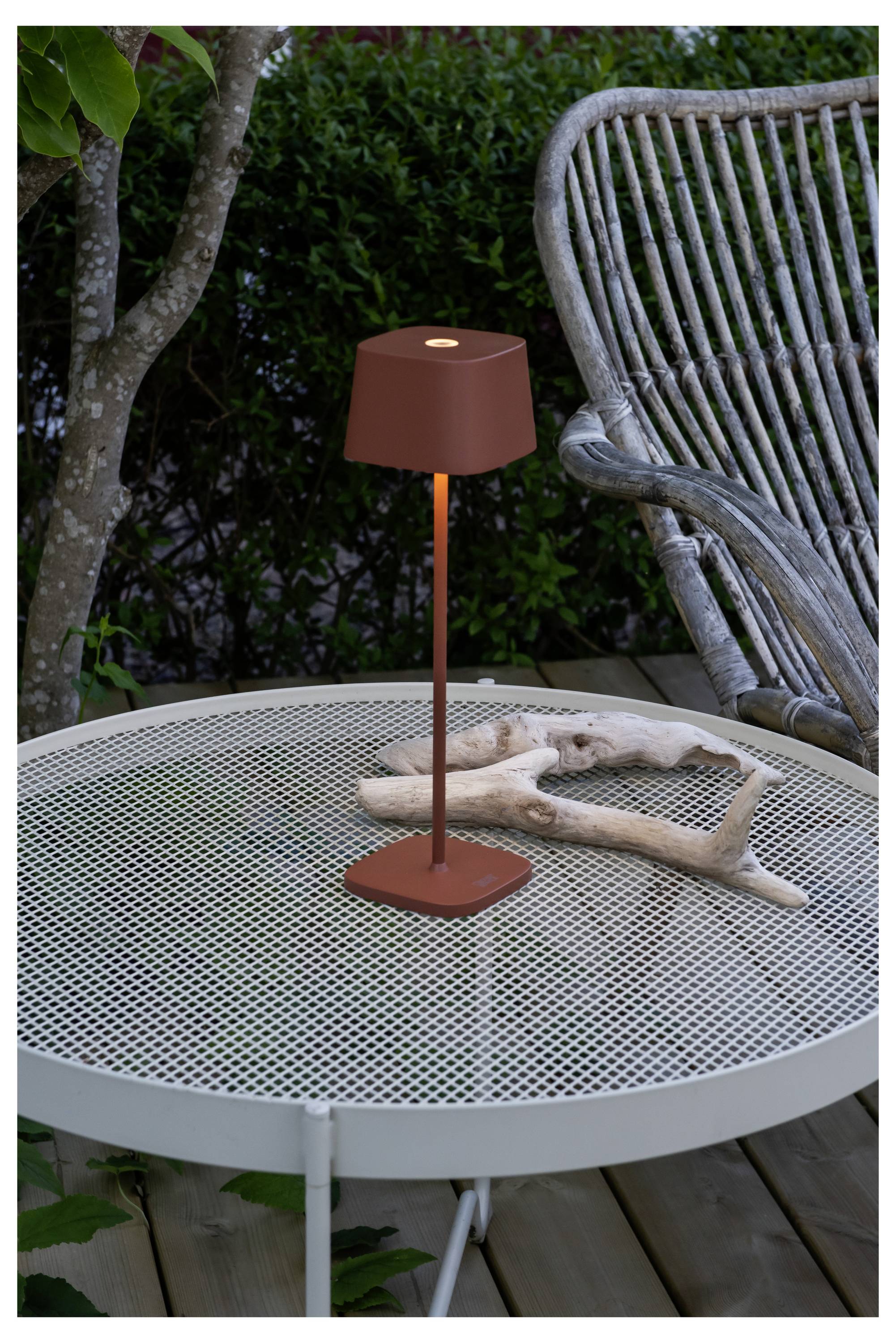 Eine kleine, rote Außenlampe auf einem weißen Maschtisch neben Treibholzdekoration; ein Korbstuhl und Grünpflanzen befinden sich im Hintergrund.