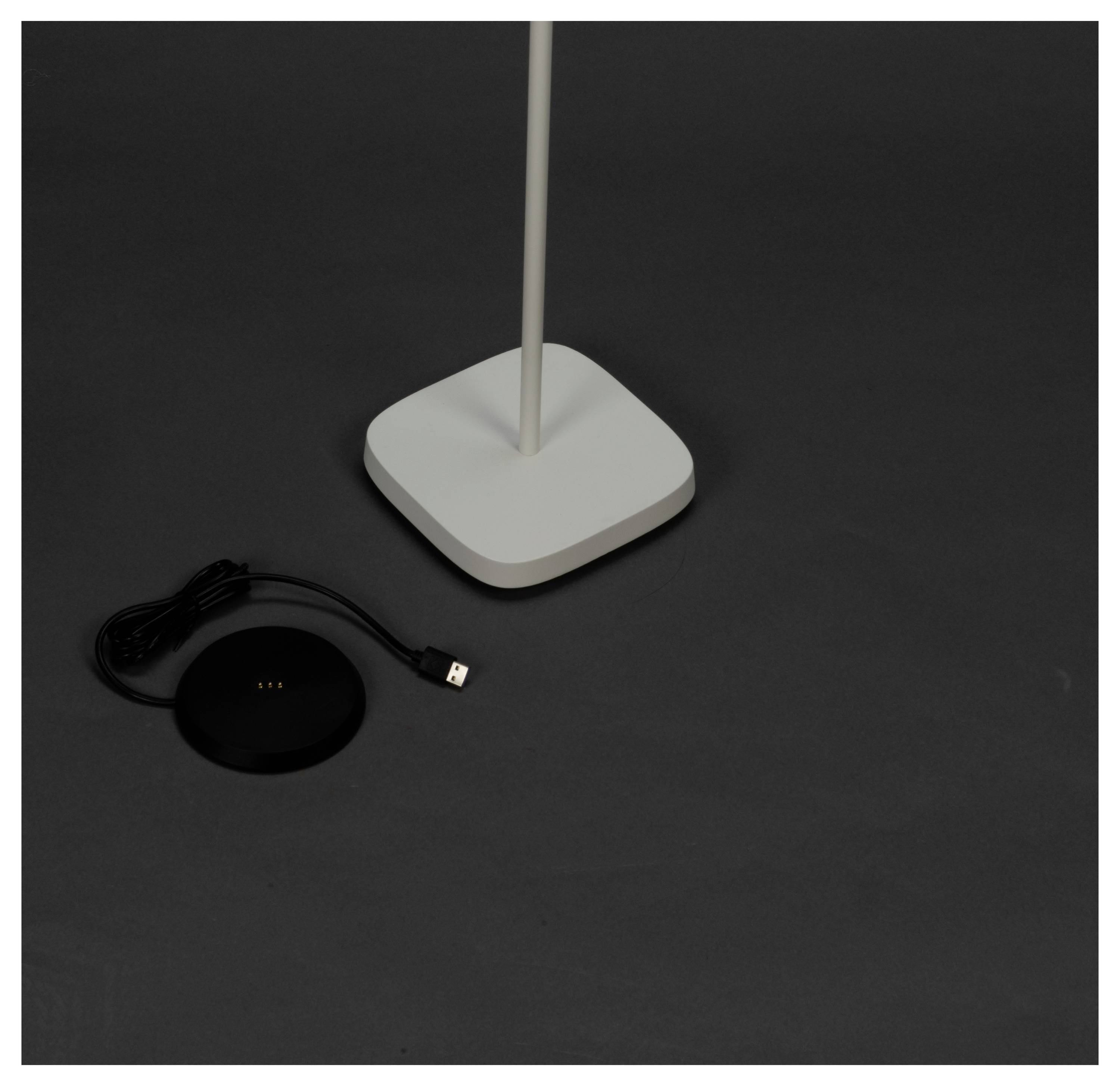 Eine weiße, quadratische Lampe steht auf einer dunklen Oberfläche, mit einem schwarzen, runden Ladegerät und einem angeschlossenen USB-Kabel in der Nähe.
