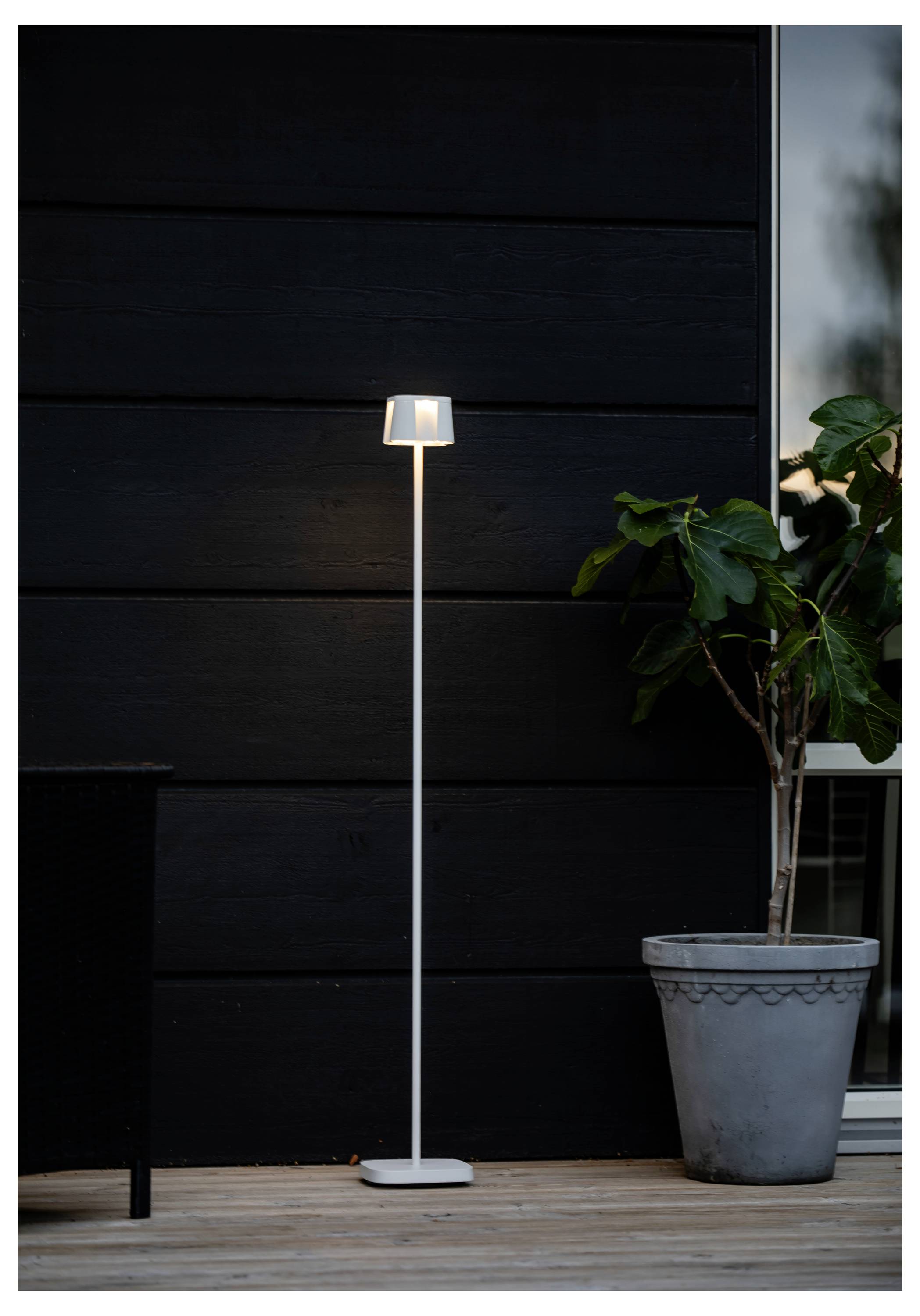 Eine hohe, schlanke Stehlampe mit einem geometrischen Lampenschirm steht beleuchtet vor einer dunklen Außenwand neben einer großen Kübelpflanze auf einer Holzterrasse.
