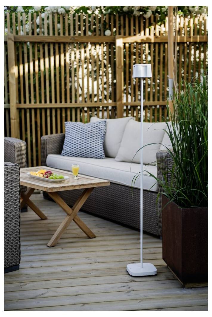 Überdachte Terrasse mit Rattansofa, Holztisch mit Snacks und Saft und hoher Stehlampe. Umgeben von Holzzaun und Grünpflanzen.