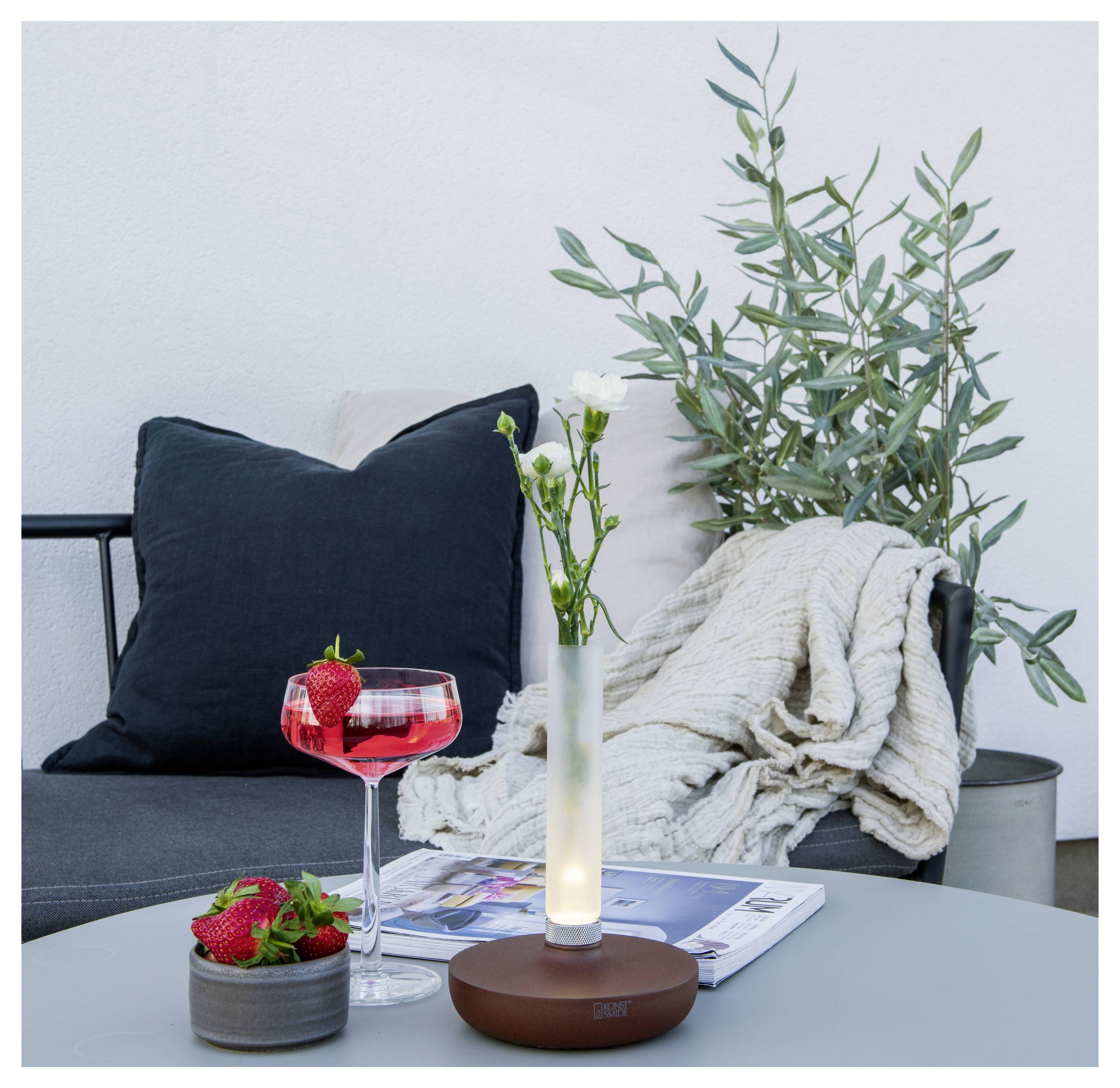 Eine gemütliche Terrasse mit einem grauen gepolsterten Stuhl, einer leichten Decke, einer Topfpflanze und einem Tisch mit einem Cocktail, Erdbeeren und einer beleuchteten Vase.