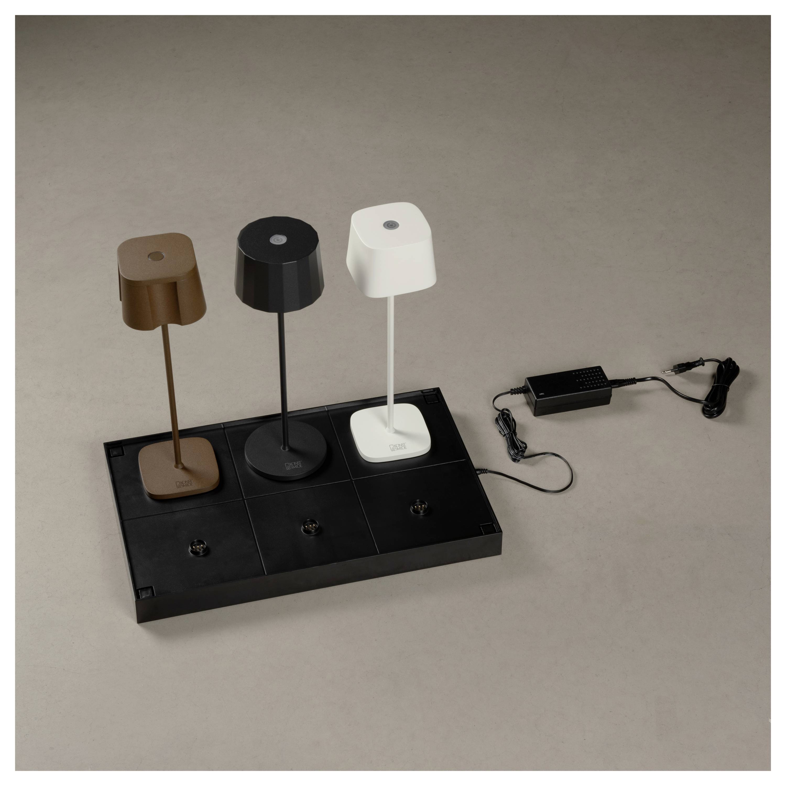 Drei moderne Tischlampen in Braun, Schwarz und Weiß sind auf einem schwarzen Wireless-Charging-Pad platziert und mit einem Netzteil verbunden.