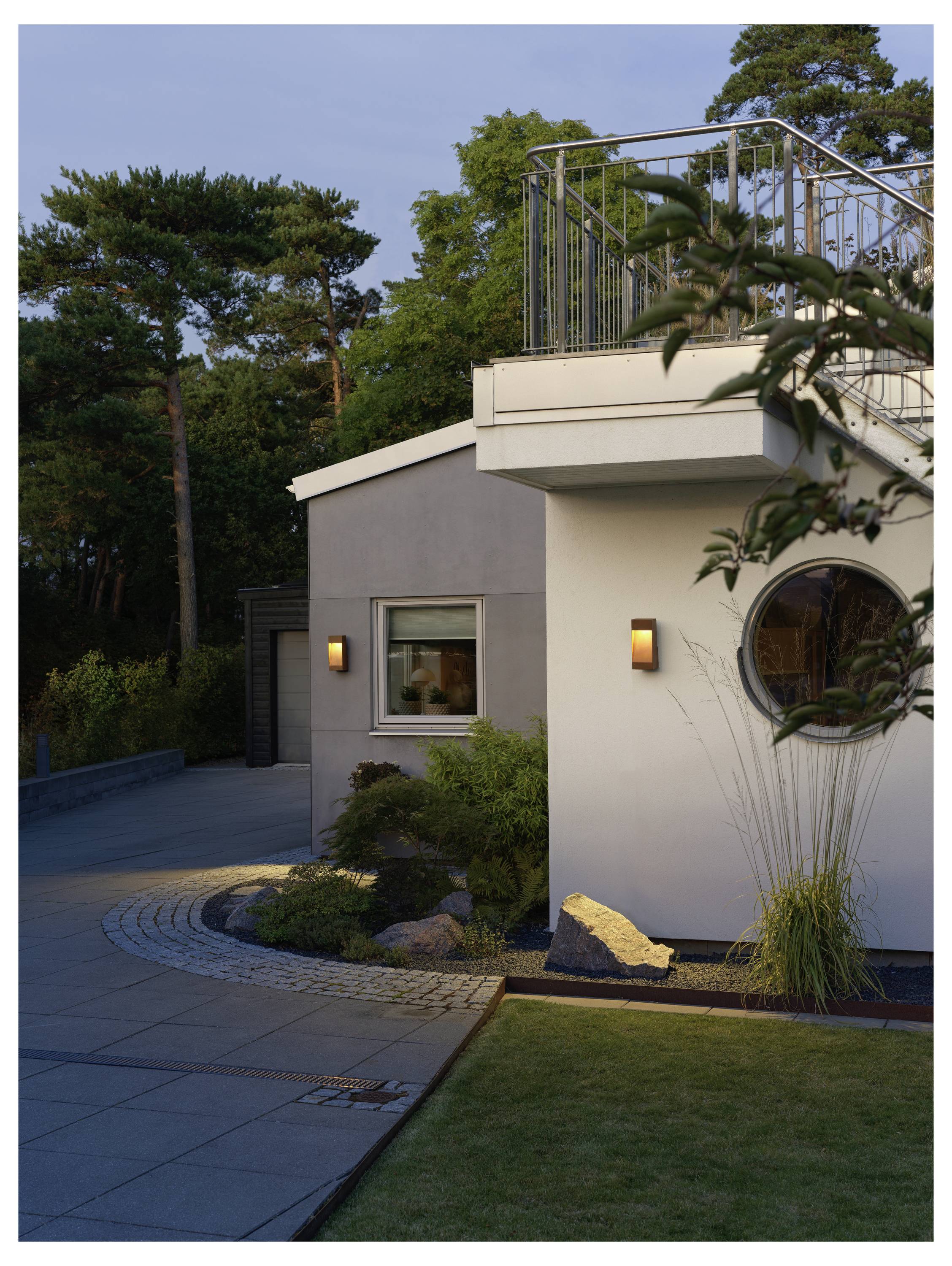 Eine moderne Hausaußenansicht in der Abenddämmerung mit beleuchteten Wandlampen, rundem Fenster und umgebender Vegetation, die eine ruhige Atmosphäre erzeugt.