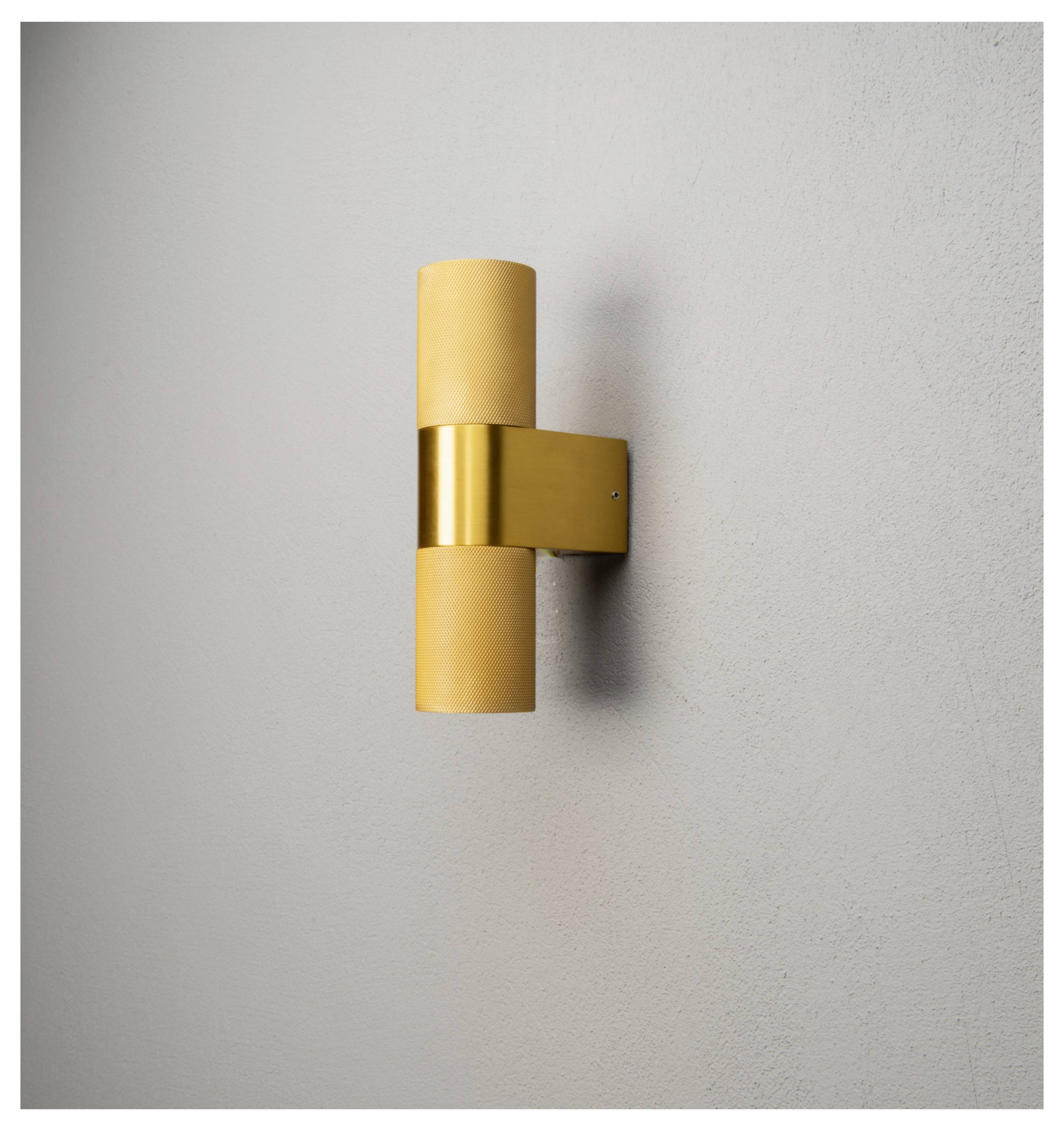 Eine minimalistische goldene Wandleuchte mit zwei zylindrischen Lampen, an einer hellgrauen Wand montiert, die modernes Ambiente-Licht erzeugt.