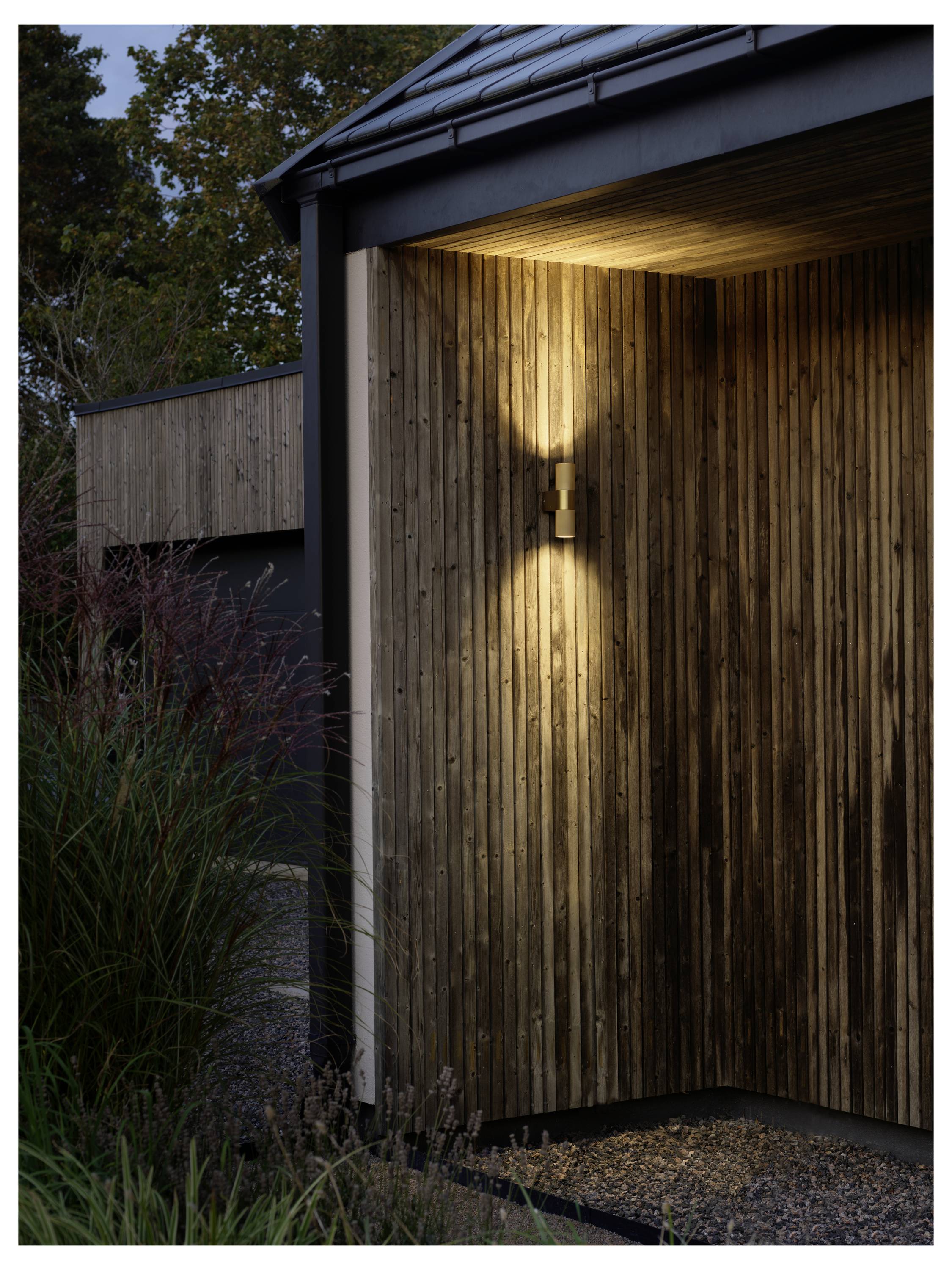 Eine Holzaußenwand mit modernem Design wird von einem warmen Licht sanft beleuchtet und befindet sich inmitten einer Gartenumgebung bei Einbruch der Dämmerung.